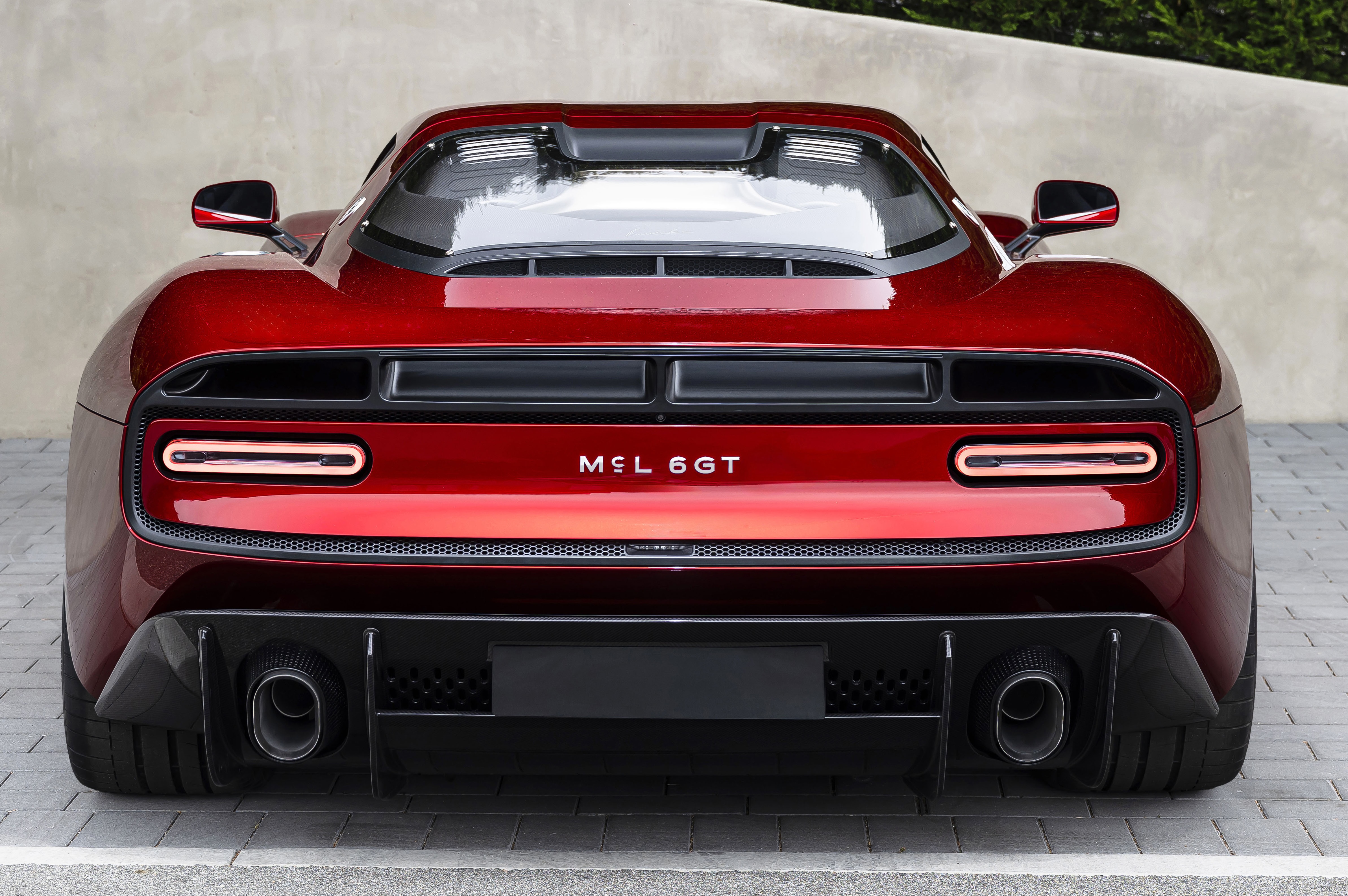 Traseira de um carro esportivo vermelho brilhante, com lanternas horizontais finas e o nome MCL 6GT no centro. Possui duas saídas de escapamento circulares e um difusor preto na parte inferior, sobre um piso de blocos cinzas