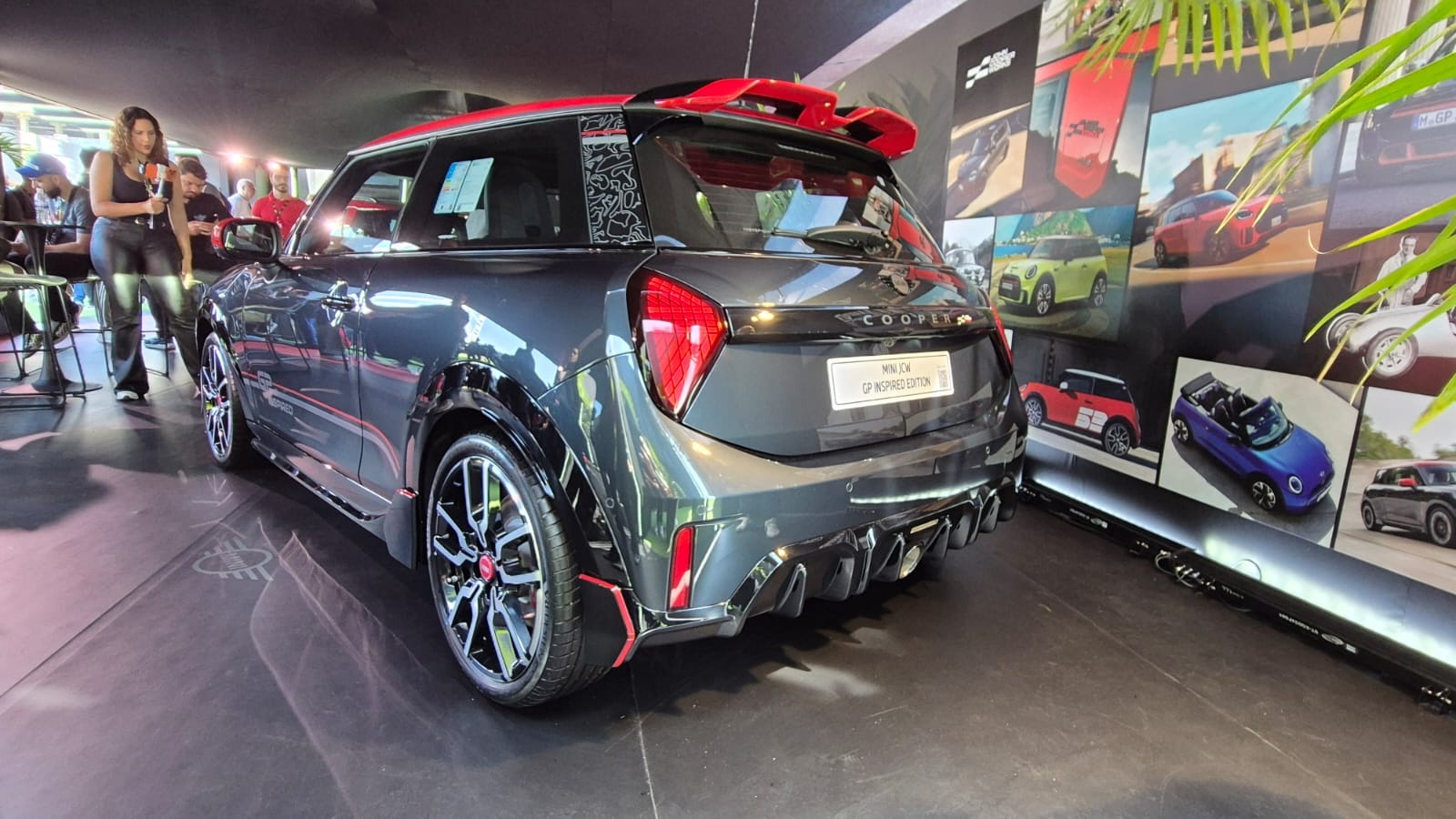 Mini Cooper cinza escuro com teto e aerofólio vermelhos, exibido em um evento. Uma mulher de calça e blusa pretas segura um microfone à esquerda. Ao fundo, uma parede com fotos de carros coloridos e folhagens verdes no canto direito