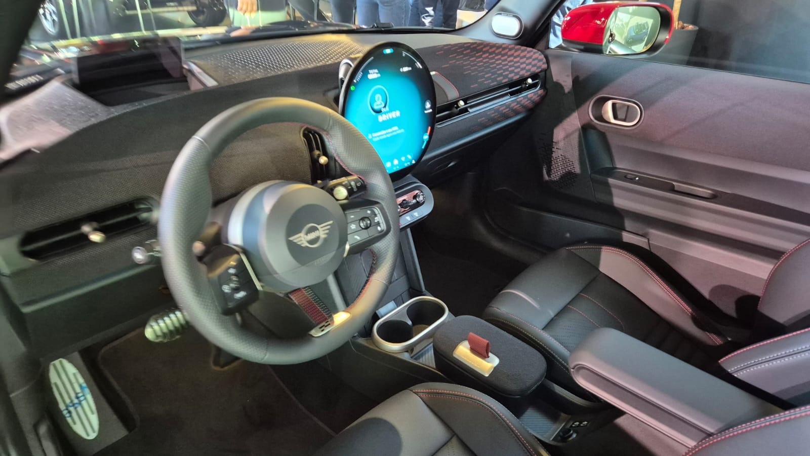Interior de um carro Mini Cooper com volante preto e o logotipo da marca no centro. Um painel digital redondo e azul exibe ícones de perfil e configurações. O banco do motorista é preto com detalhes em vermelho, e o console central tem porta-copos e um apoio de braço. Um retrovisor vermelho é visível à direita