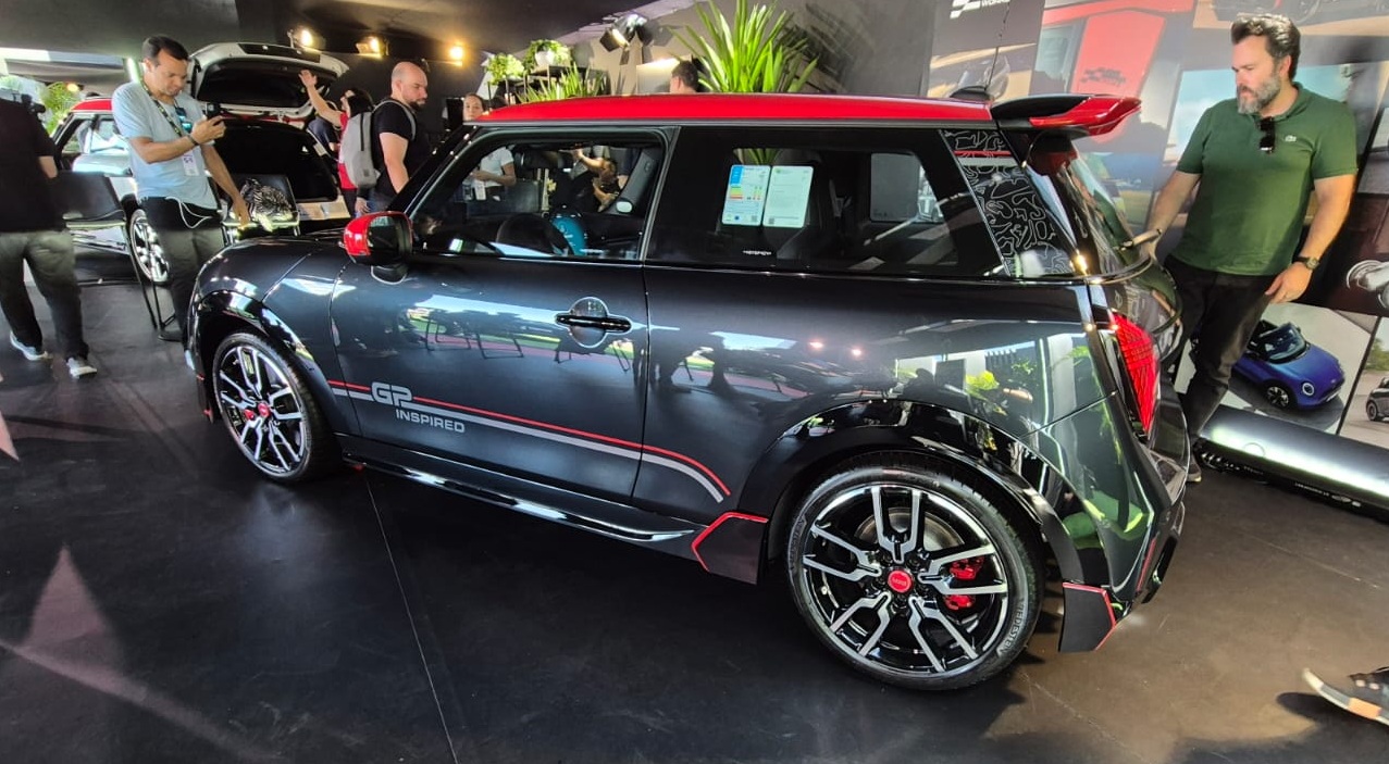 Mini Cooper cinza escuro com teto vermelho e detalhes vermelhos, exposto em um evento. Homens observam o carro e outros veículos ao fundo