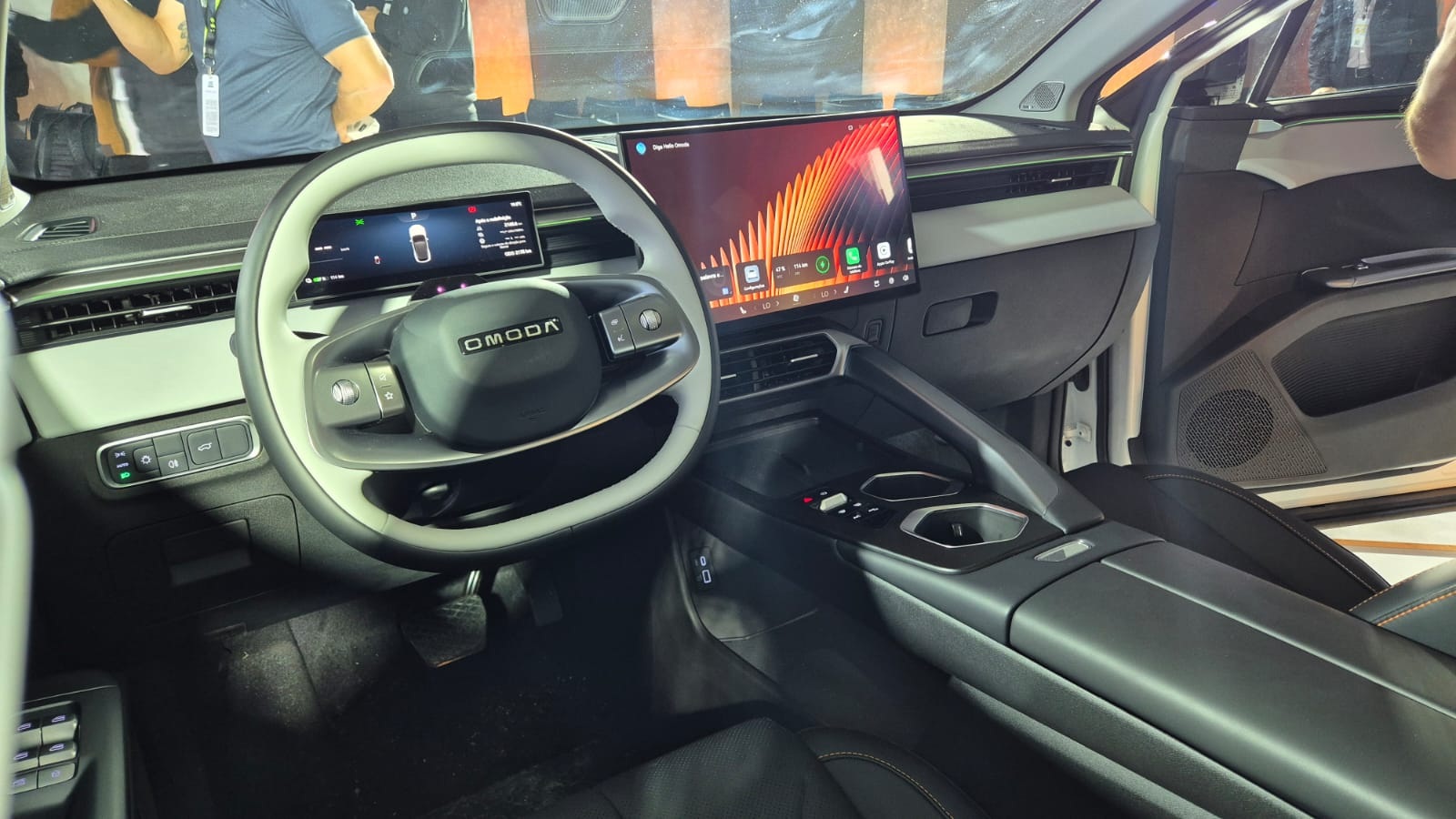 Interior de um carro OMODA com volante multifuncional cinza e branco, painel digital e tela multimídia grande exibindo aplicativos. O console central tem porta-copos e acabamento escuro, com detalhes em cinza claro no painel e portas.
