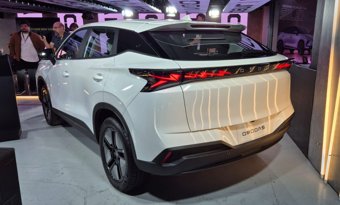 SUV OMODAS branco, visto de traseira e lateral direita, com lanternas vermelhas acesas e o nome OMODAS no porta-malas. Pessoas e outro carro desfocados ao fundo, em um ambiente de exposição