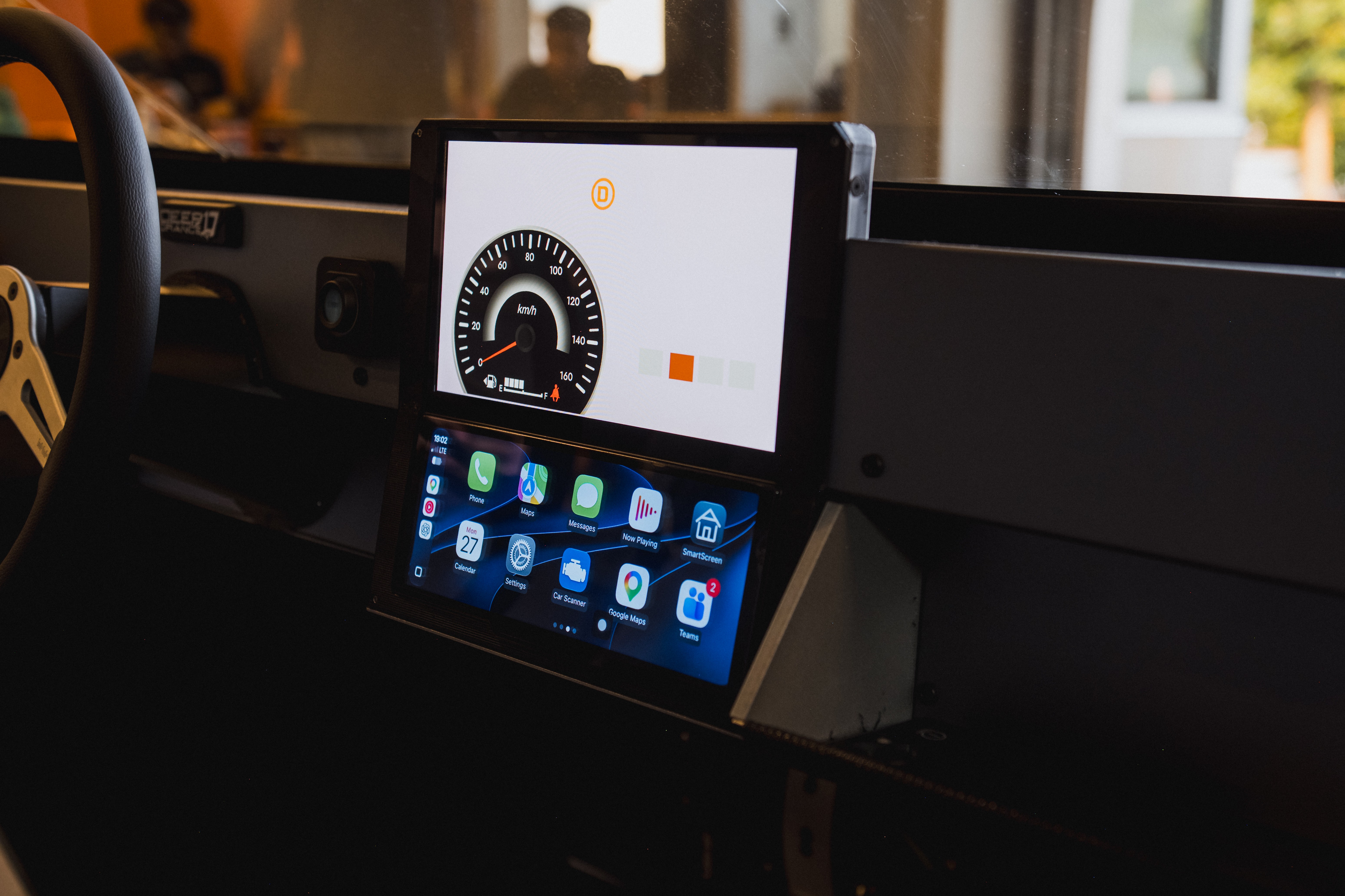 Painel de carro com dois monitores digitais. O superior exibe um velocímetro e o inferior, aplicativos de celular em ícones coloridos. Volante preto à esquerda.