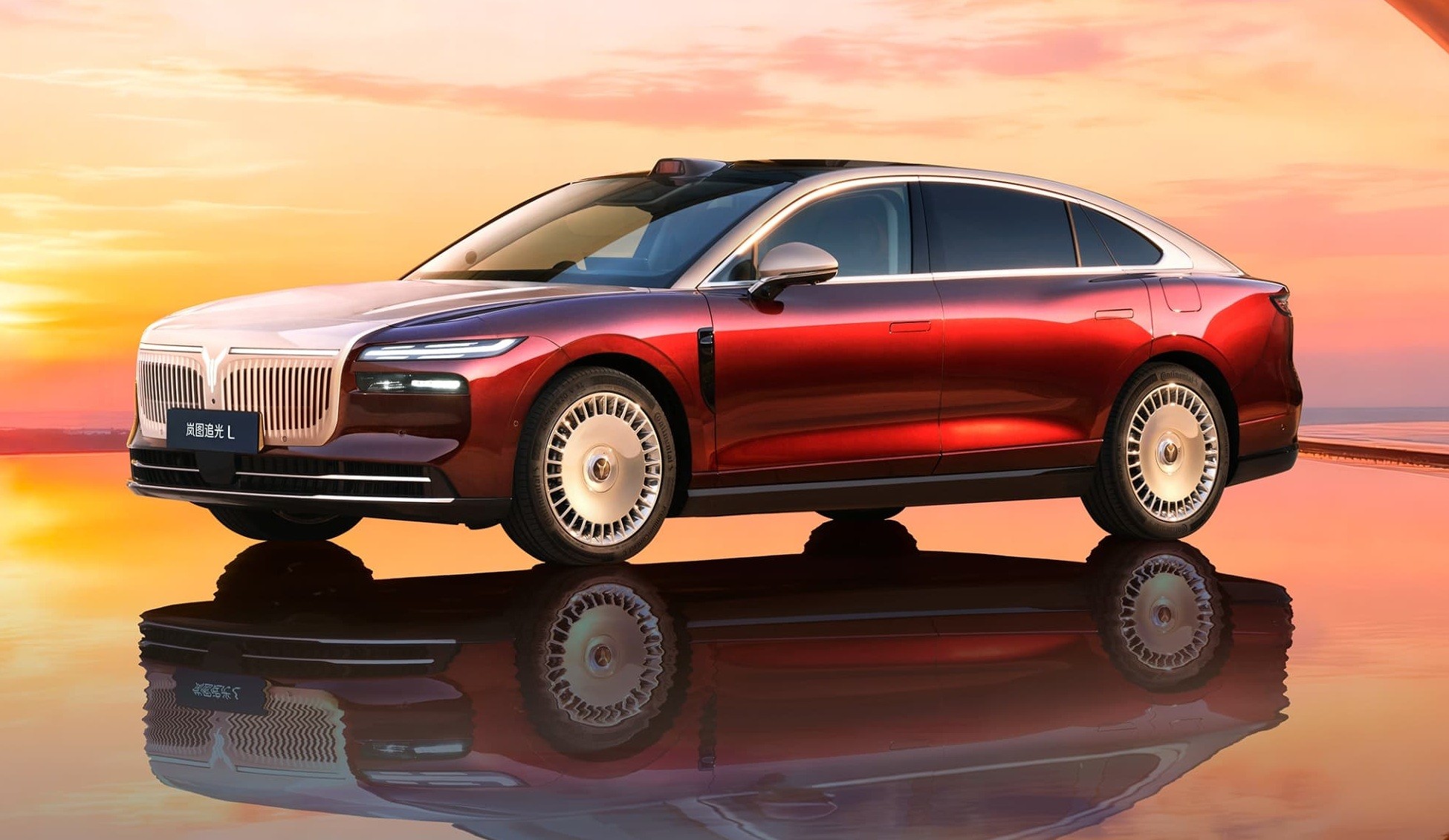 Carro de luxo vermelho e branco com teto preto, parado sobre superfície espelhada que reflete sua imagem, contra um céu alaranjado de pôr do sol