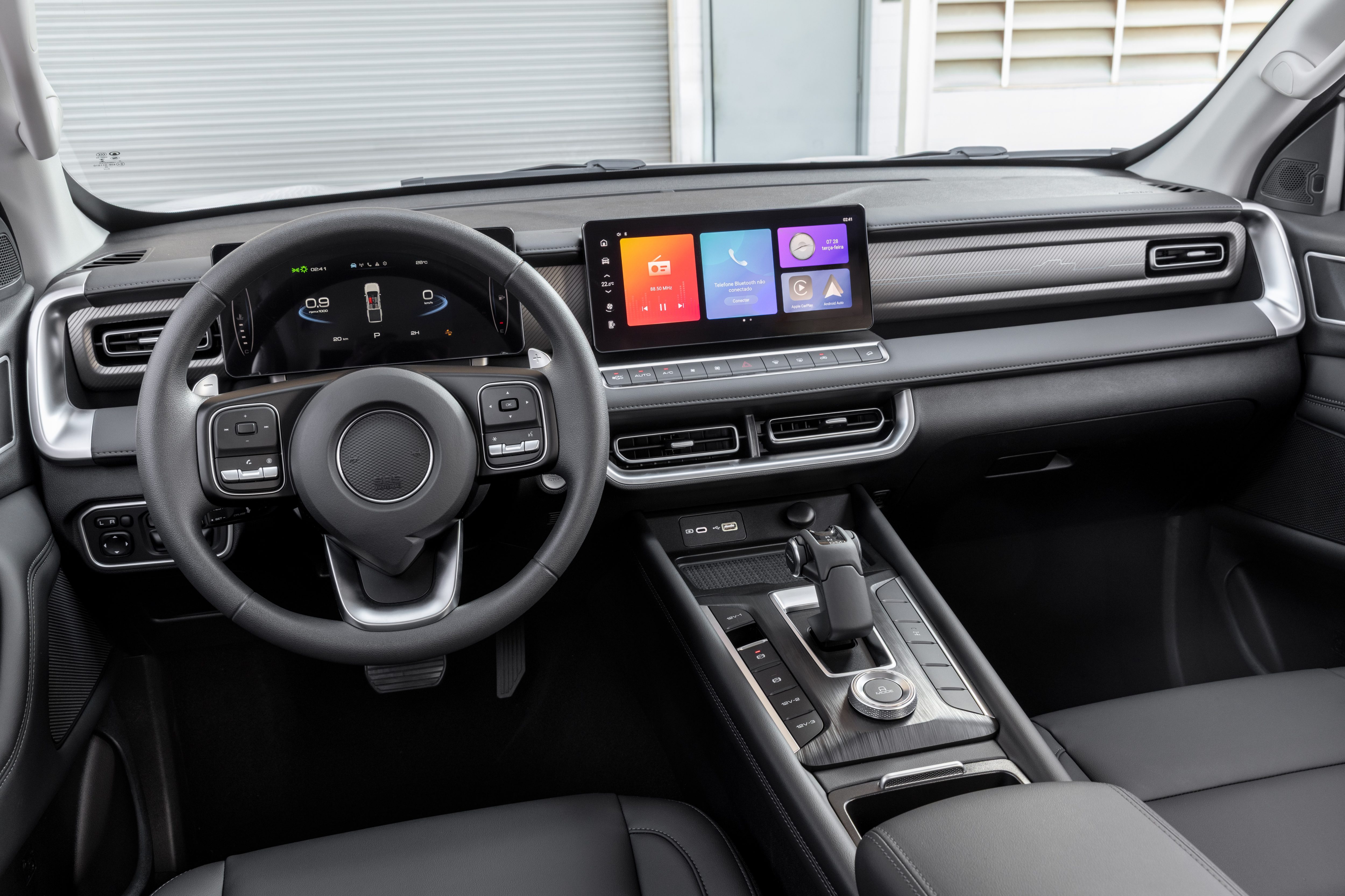 Interior de um carro moderno com painel preto e detalhes prateados. Volante multifuncional, painel digital e tela multimídia central com ícones coloridos. Câmbio automático e console central com botões e acabamento em madeira escura. Bancos de couro pretos visíveis na parte inferior.