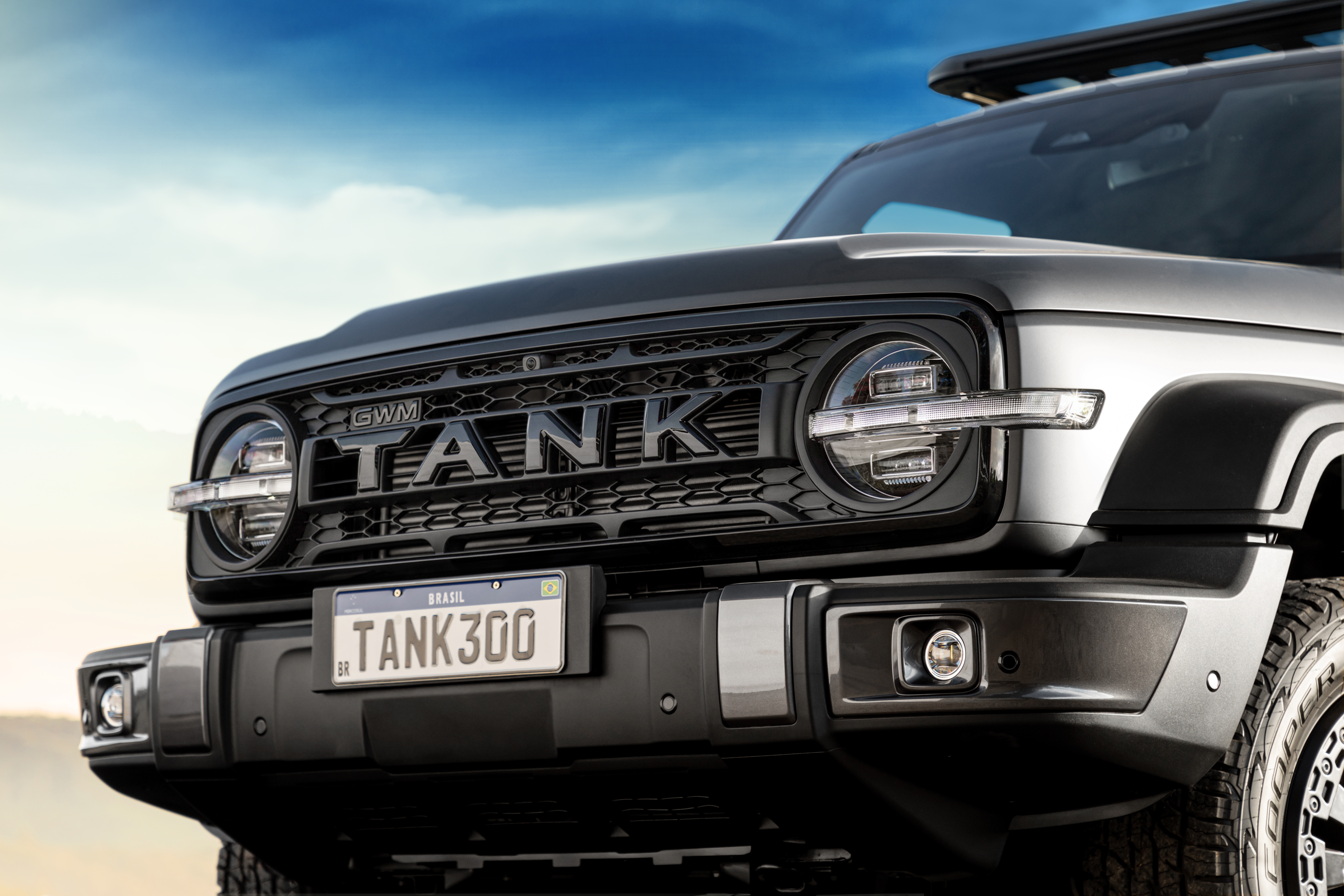 Parte frontal de um SUV GWM Tank 300 cinza, com grade preta e faróis redondos acesos. A placa de carro brasileira exibe TANK300