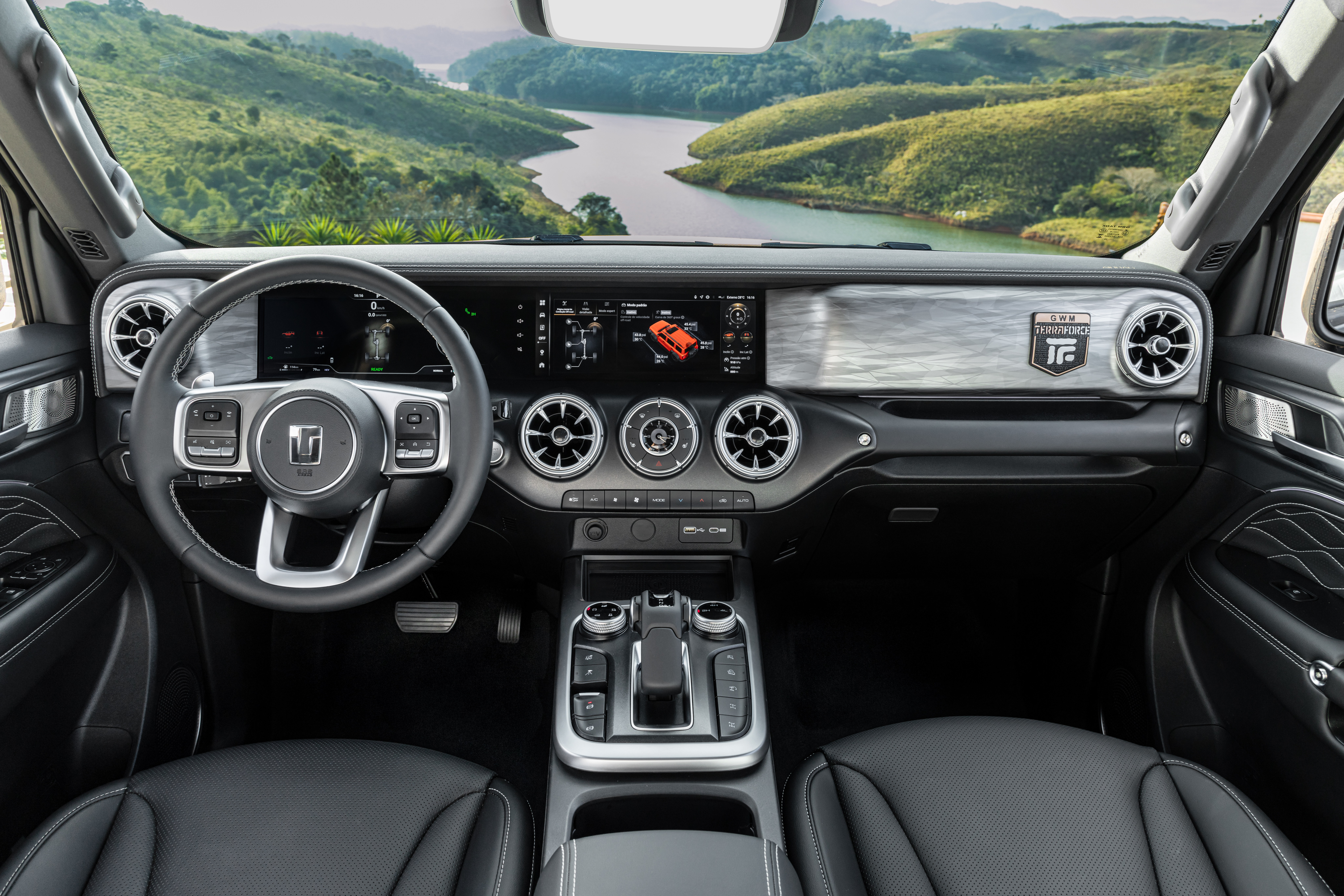 Interior de um carro moderno com volante multifuncional, painel digital exibindo um veículo vermelho e saídas de ar circulares. O console central possui câmbio automático e botões de controle. Ao fundo, uma paisagem com montanhas verdes e um rio sinuoso sob um céu claro.