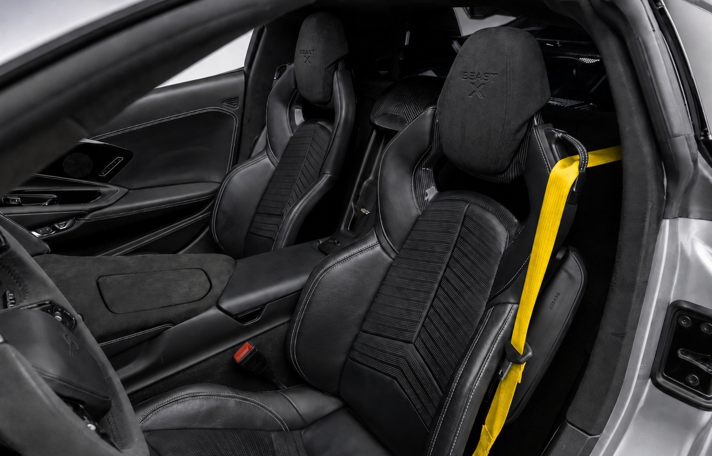 Interior de um carro esportivo com dois bancos dianteiros pretos de couro e tecido, costuras brancas, e o logotipo BEAST X nos encostos de cabeça. O cinto de segurança do passageiro é amarelo vibrante, contrastando com o ambiente escuro. O console central e o painel da porta também são pretos, com detalhes em costura branca