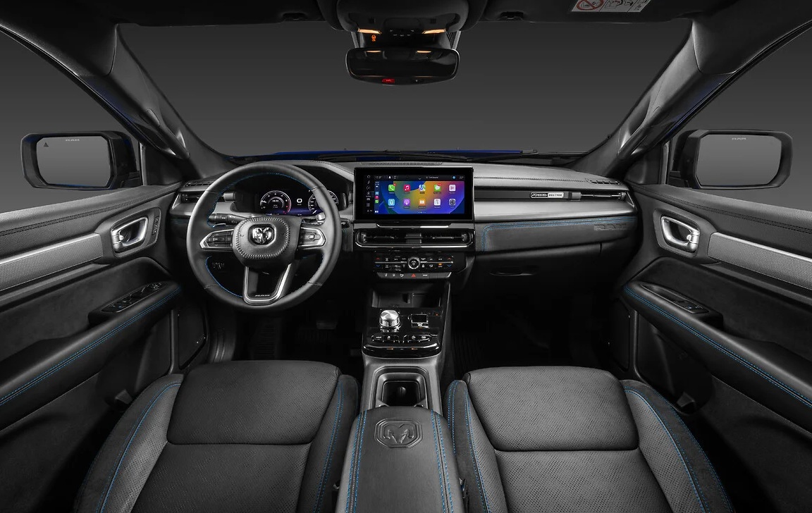 Interior de um carro moderno com bancos de couro escuro e costura azul, volante multifuncional com logo Ram, painel digital e central multimídia com tela sensível ao toque exibindo ícones coloridos
