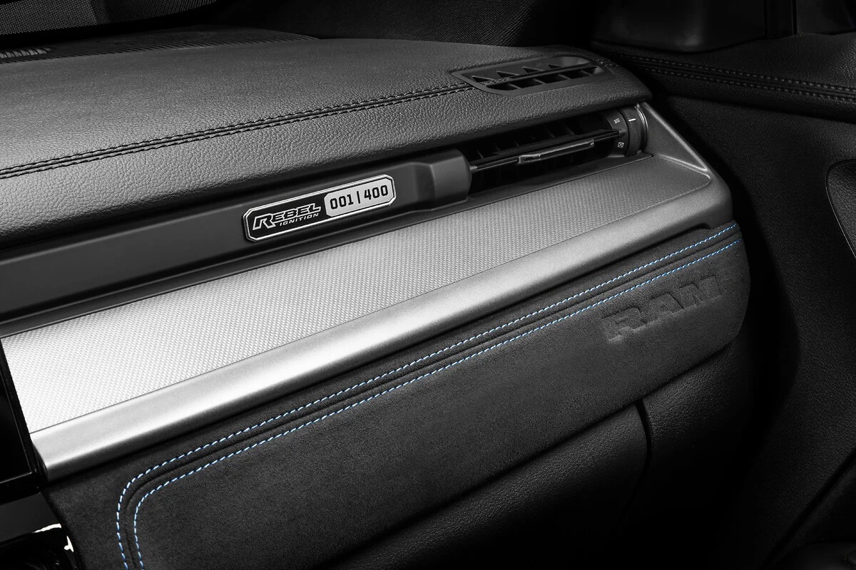 Detalhe do painel de um carro, com acabamento em couro preto e costura azul. Uma placa metálica com REBEL IGNITION EDITION 001/400 está visível, acima de uma faixa prateada texturizada. O logotipo RAM está gravado no couro abaixo