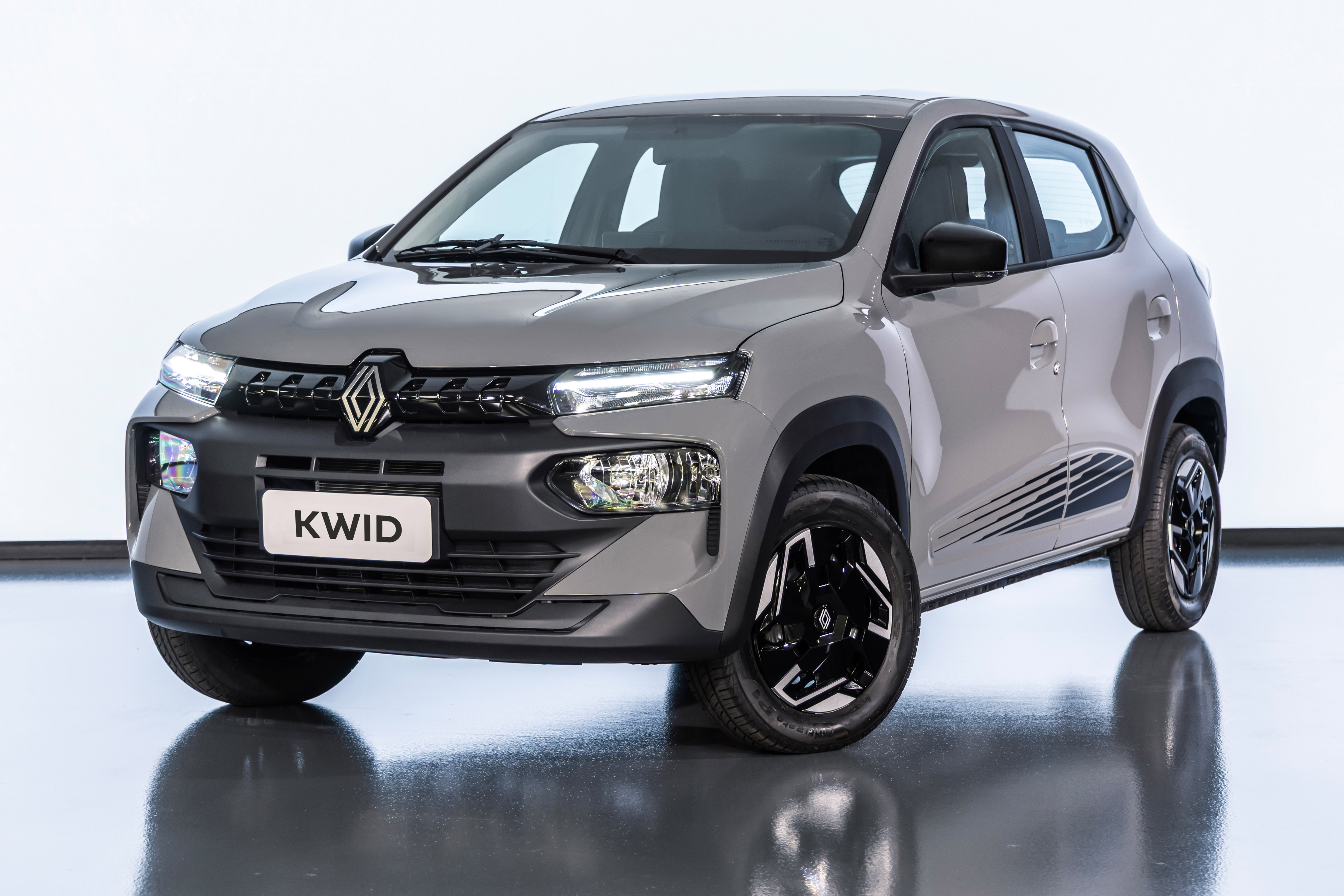 Carro Renault Kwid cinza claro, visto de frente e ligeiramente à direita, com faróis acesos, grade frontal escura e placa KWID branca. Rodas pretas com detalhes brancos e faixas decorativas pretas na lateral inferior, sobre piso reflexivo e fundo branco