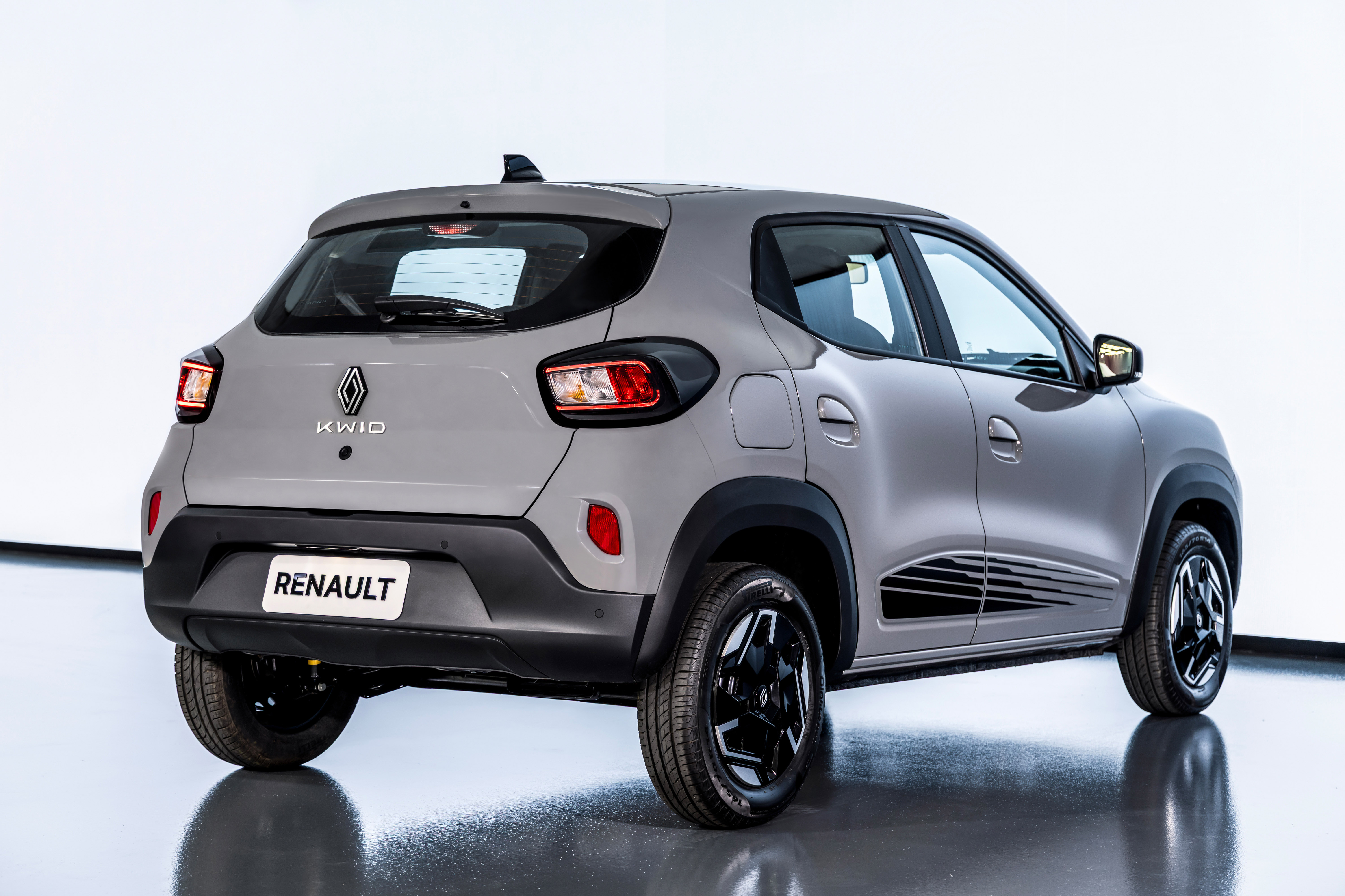 Traseira e lateral direita de um carro Renault Kwid cinza claro, com detalhes em preto nos para-lamas e na parte inferior da porta, sobre um piso escuro e reflexivo, com fundo branco