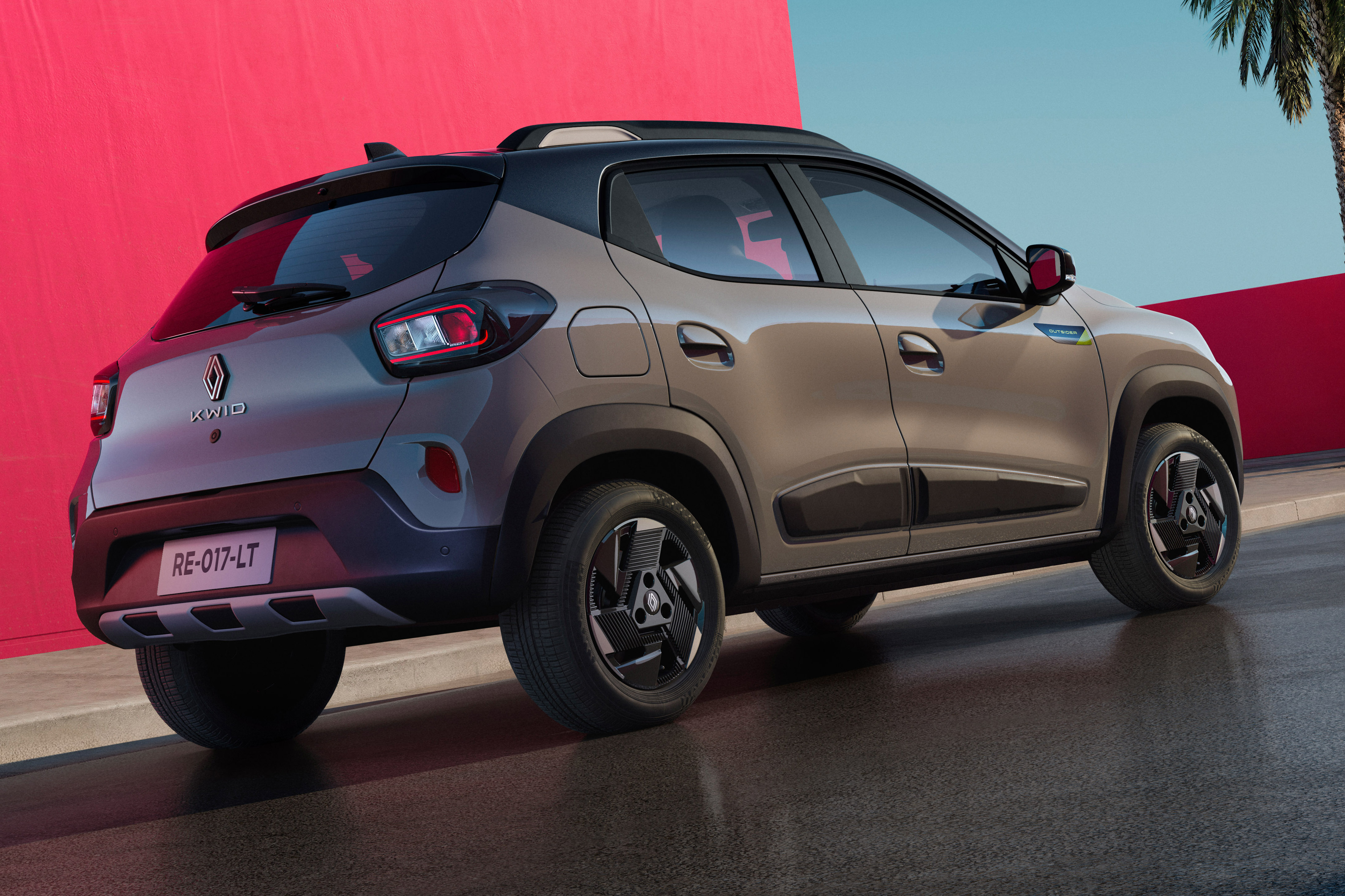 Carro cinza-claro, modelo SUV compacto, visto de traseira e lateral, com teto preto e detalhes pretos nas caixas de roda. O veículo está parado em uma rua escura, com um muro vermelho vibrante ao fundo e um céu azul-claro com uma palmeira à direita
