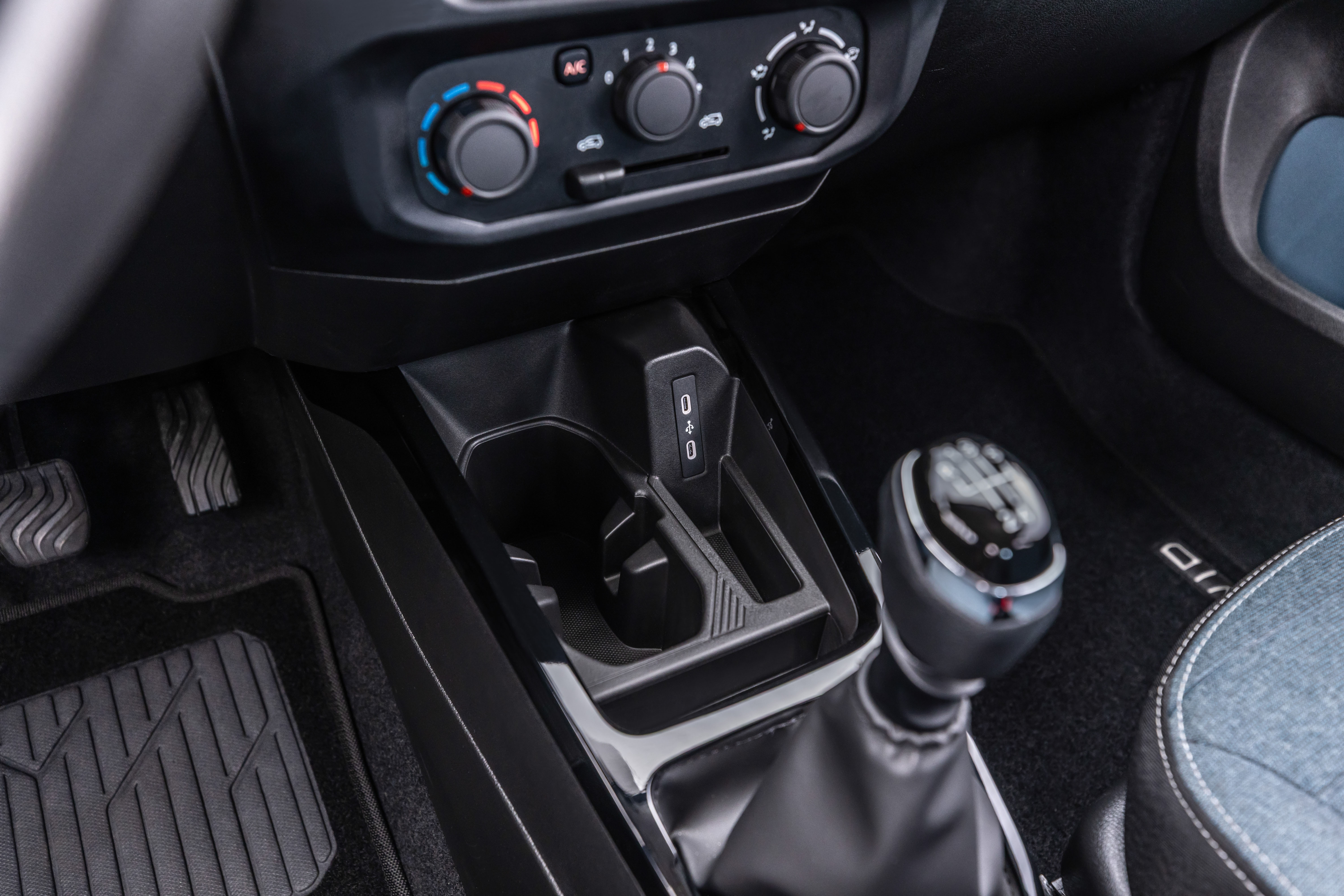 Interior de carro com câmbio manual de cinco marchas em primeiro plano, porta-copos e duas entradas USB no console central. Acima, três botões giratórios do ar-condicionado. À esquerda, pedais de freio e embreagem. Tapetes pretos no chão.