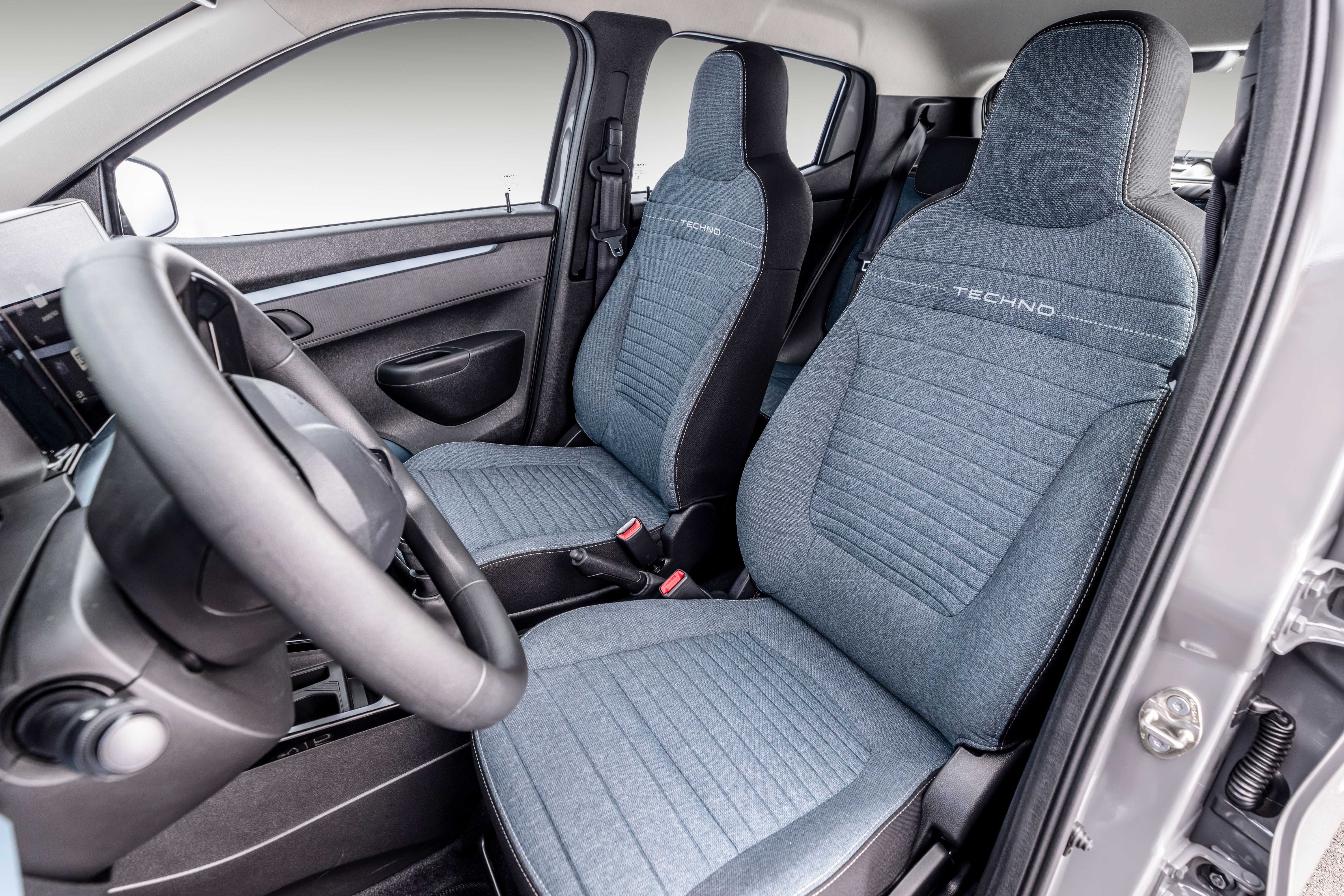 Interior de um carro com bancos dianteiros em tecido azul-acinzentado e detalhes em preto, com a palavra TECHNO bordada no encosto. O volante preto está em primeiro plano à esquerda, e a porta do passageiro está aberta à direita, revelando a dobradiça. O ambiente é claro, com luz natural entrando pelas janelas