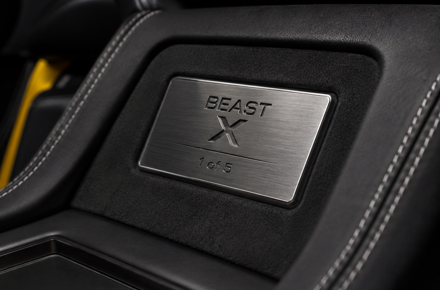 Placa metálica escovada com a inscrição BEAST X 1 of 5 em relevo, embutida no console central de um carro, cercada por estofamento preto com costura branca