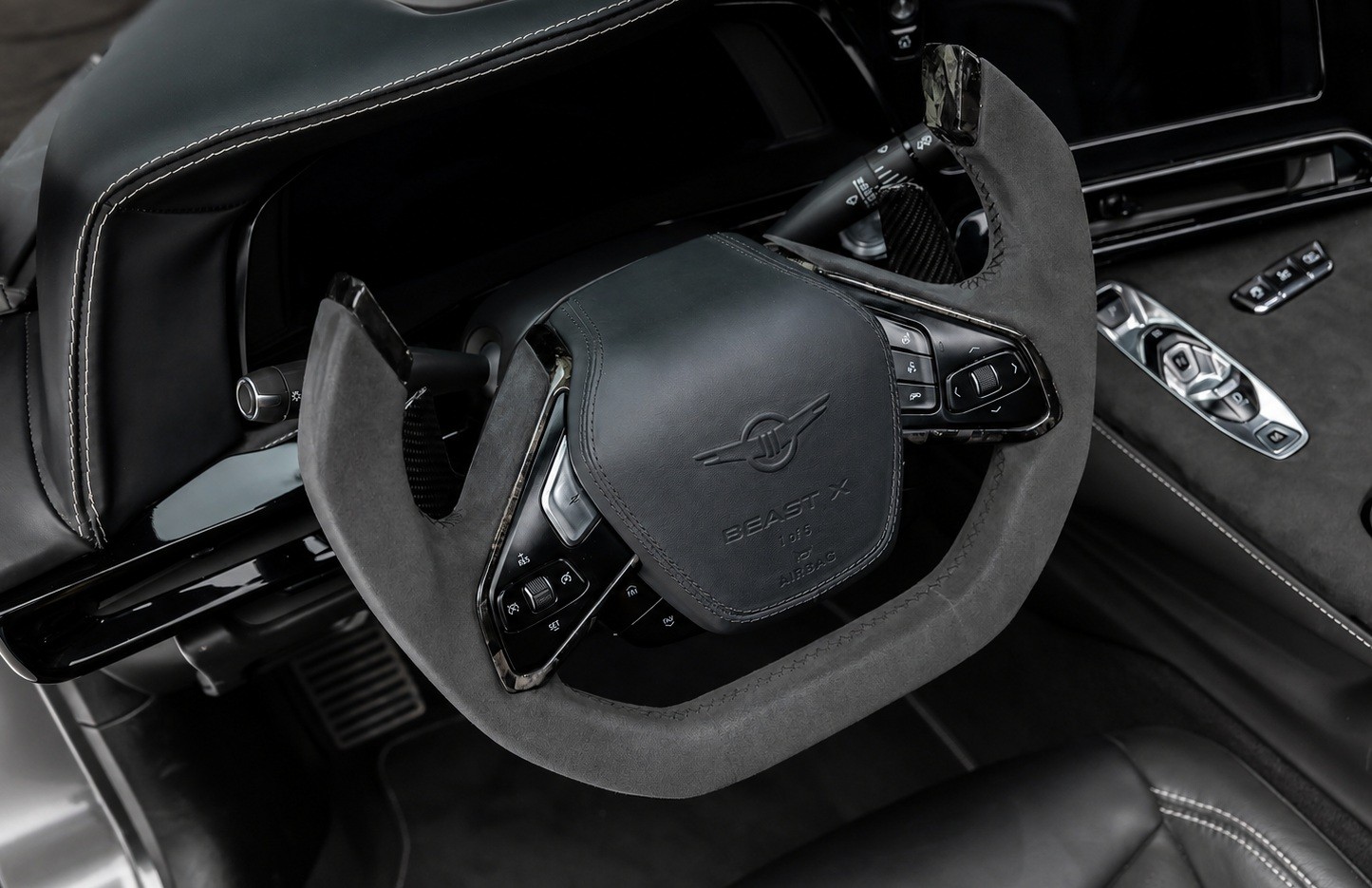 Interior de carro esportivo com volante revestido em camurça escura, costura branca e emblema Beast X no centro. À direita, painel com botões de controle e um controle remoto prateado. O ambiente é luxuoso e moderno, com detalhes em preto e cinza.