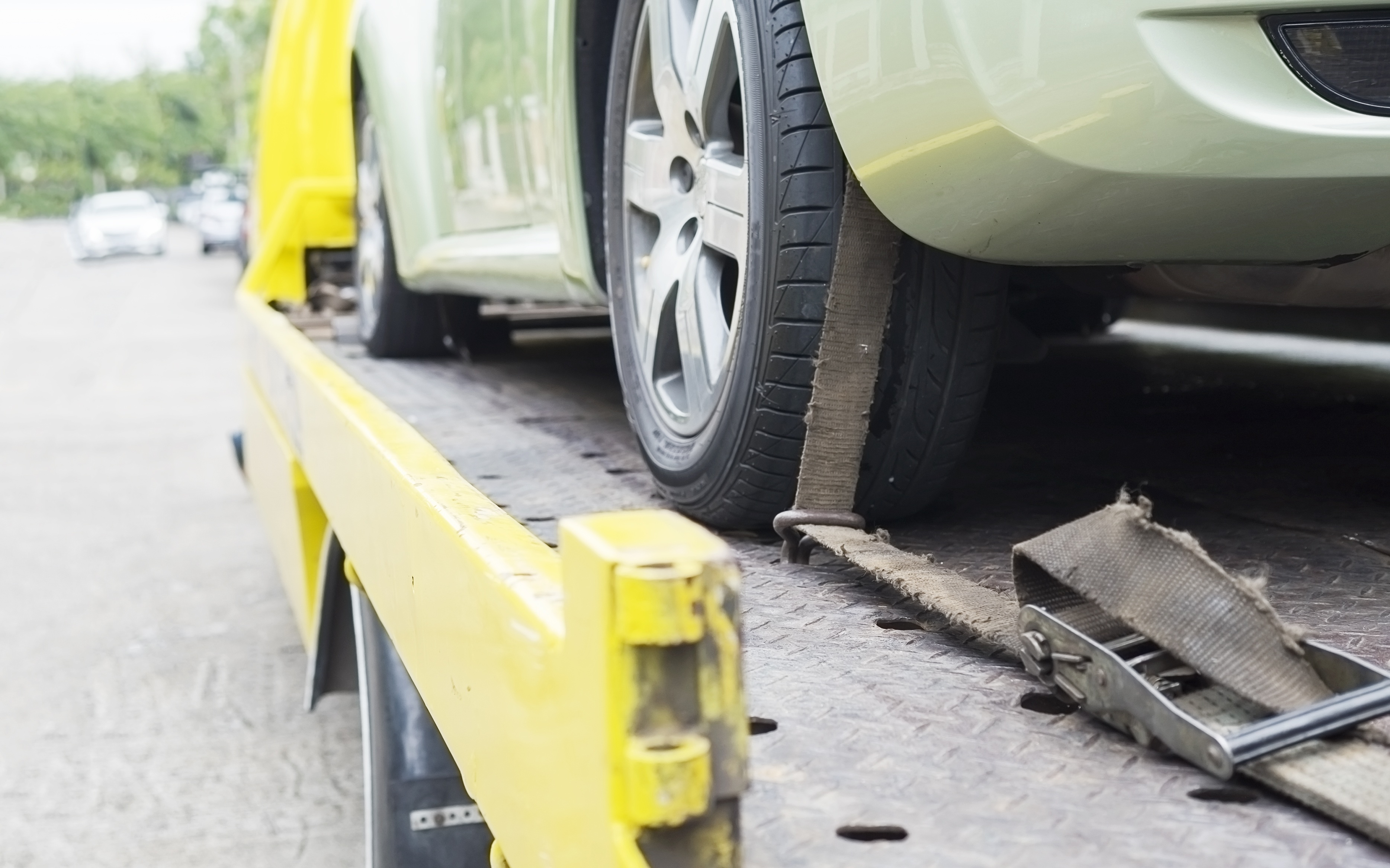 Um carro verde claro sendo transportado em um guincho amarelo, com a roda traseira presa por uma cinta de amarração desgastada. O guincho tem uma plataforma metálica texturizada e, ao fundo, outros carros desfocados em uma rua.
