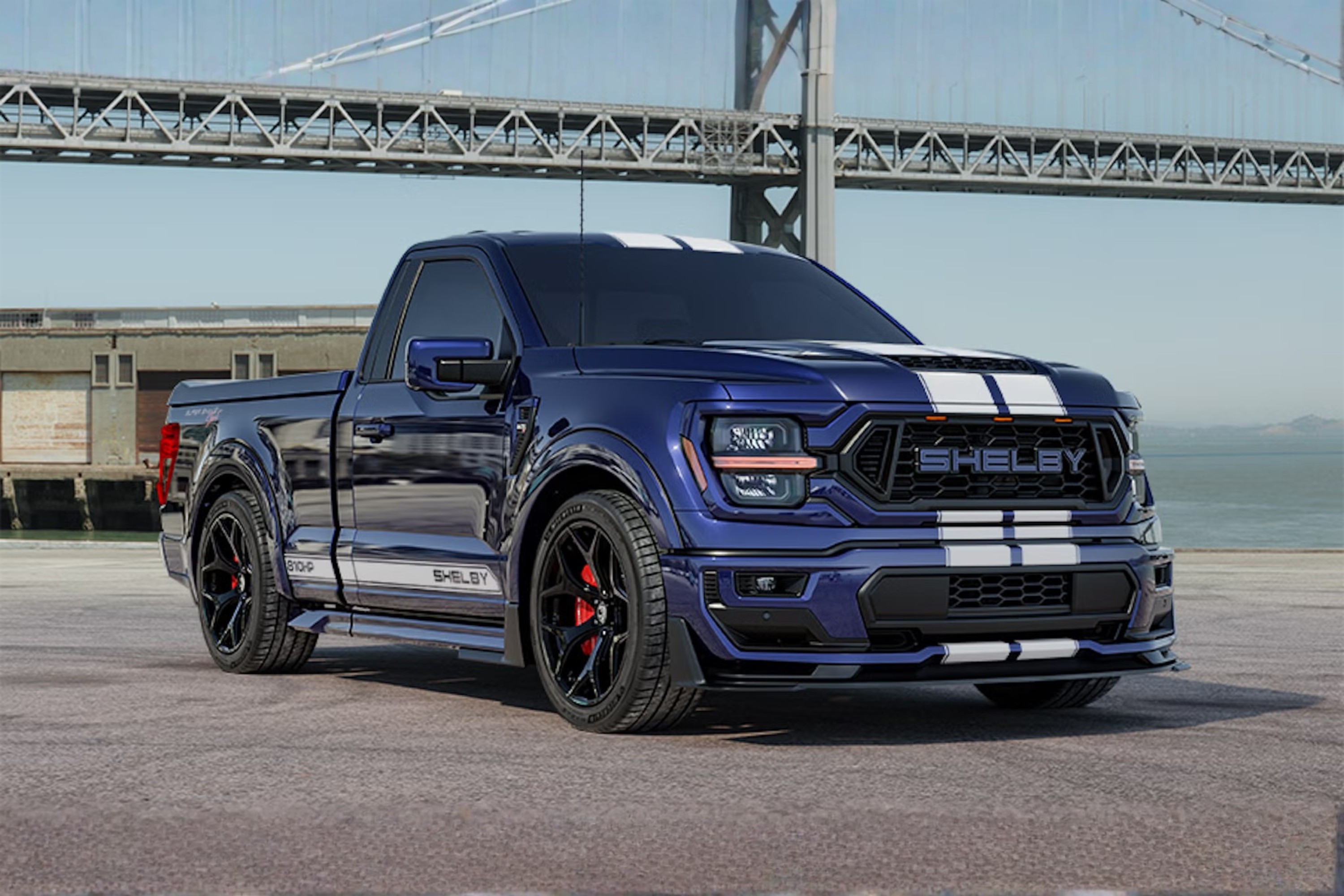 Picape Shelby F-150 azul-marinho com faixas brancas duplas no capô e teto, estacionada em um píer. Ao fundo, uma ponte metálica e o mar sob céu claro