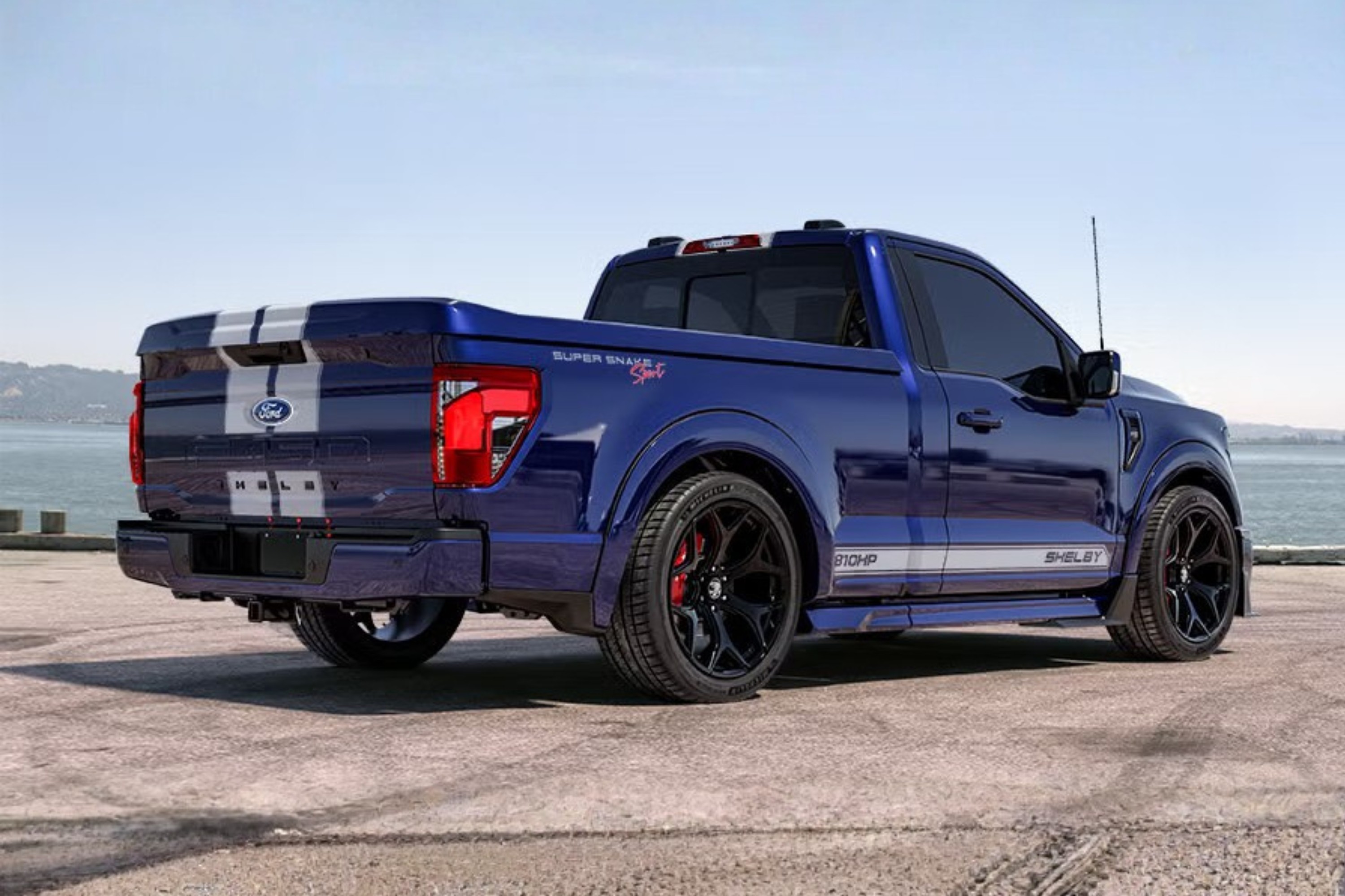 Picape Ford Shelby Super Snake Sport azul escura, vista de traseira e lateral, com faixas brancas no capô e tampa da caçamba, rodas pretas e pinças de freio vermelhas. O veículo está estacionado em um piso de asfalto, com um corpo dágua e montanhas ao fundo, sob céu claro