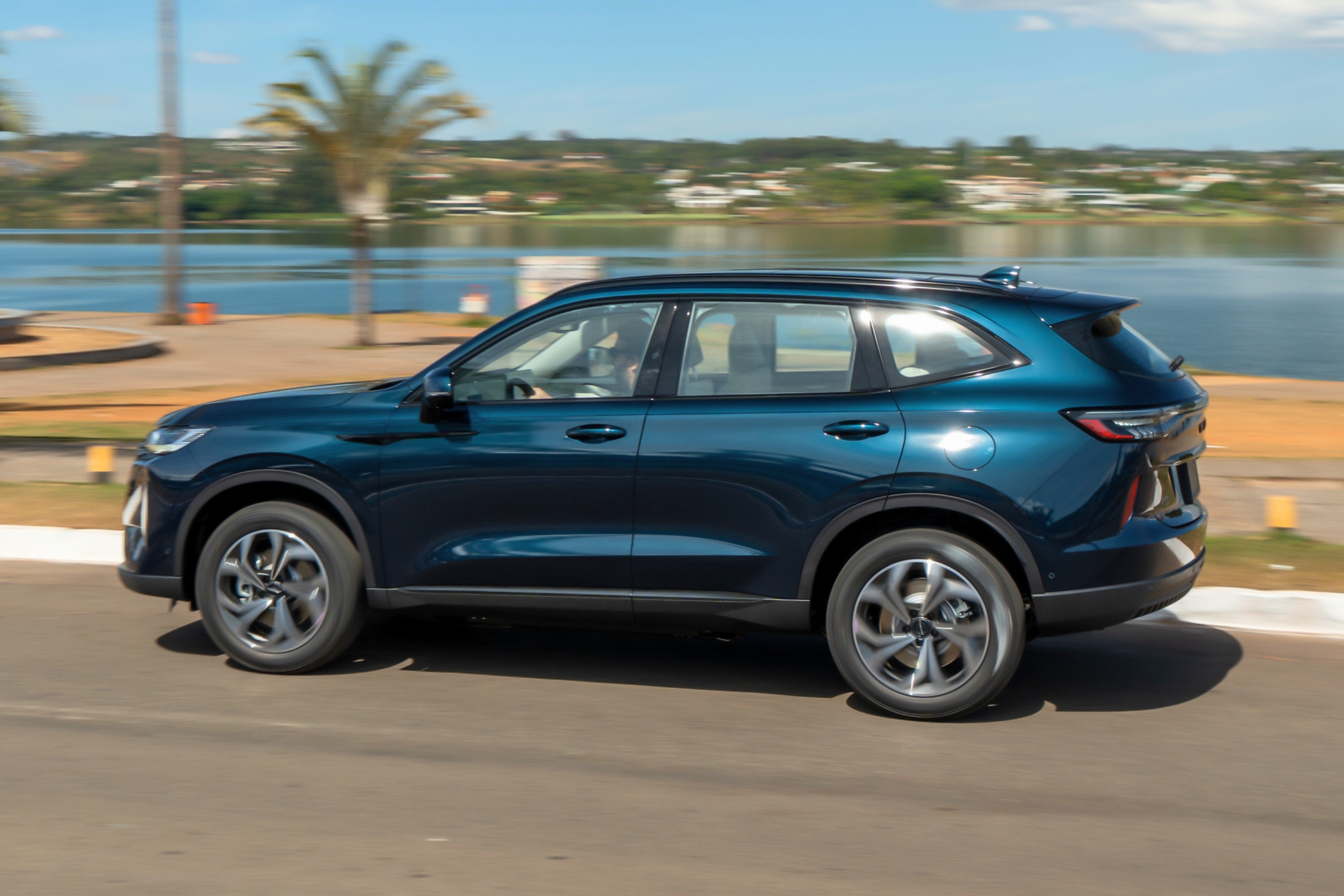 SUV azul-escuro em movimento, visto de perfil, com reflexos de luz solar na lataria. Ao fundo, um lago azul, vegetação e céu claro com nuvens esparsas