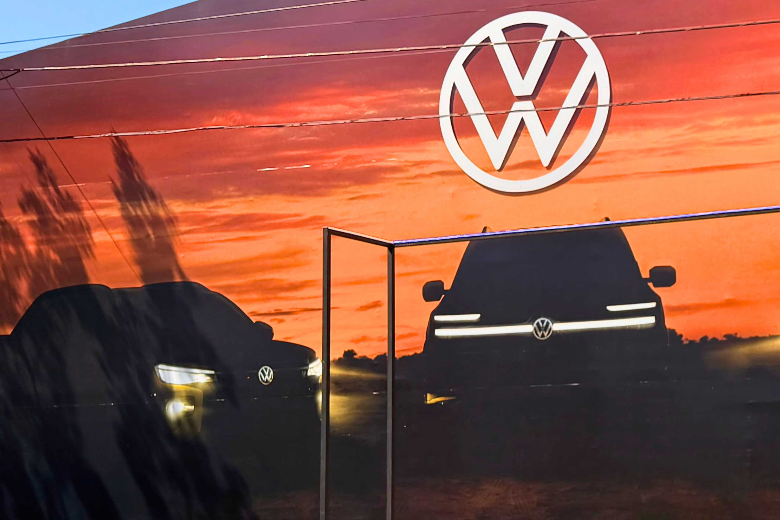 Mural da Volkswagen com o logo branco da marca no topo, sobre um p&ocirc;r do sol alaranjado. Abaixo, duas silhuetas escuras de carros com far&oacute;is acesos, um &agrave; esquerda e outro &agrave; direita, separados por uma estrutura met&aacute;lica vertical