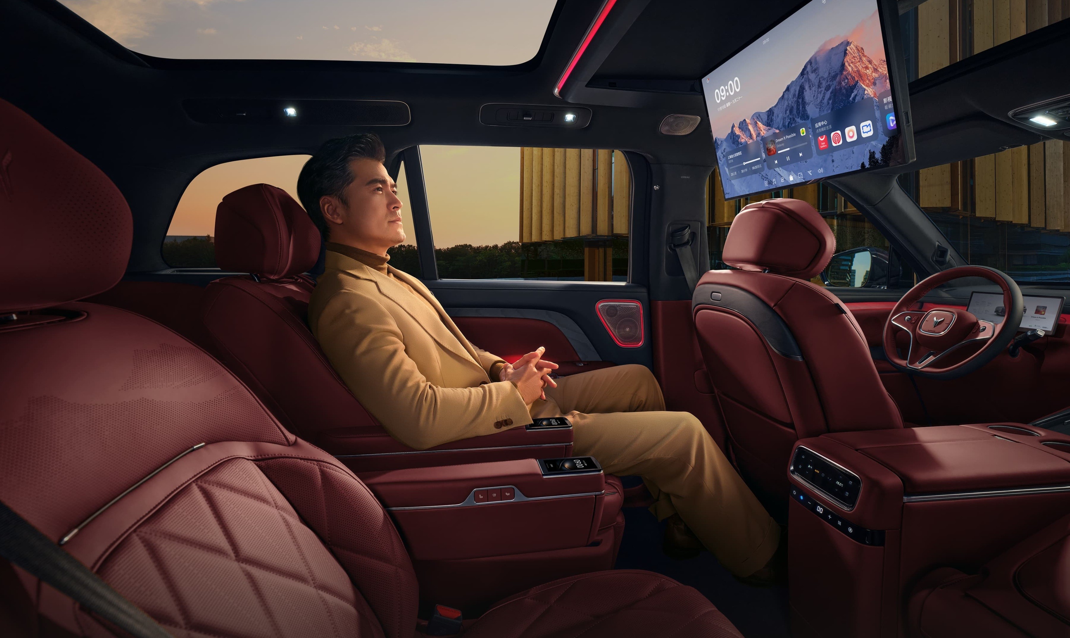 Homem asiático de terno bege relaxa no banco traseiro de carro luxuoso com estofamento vermelho, olhando pela janela. Um monitor grande exibe uma paisagem montanhosa no teto do veículo