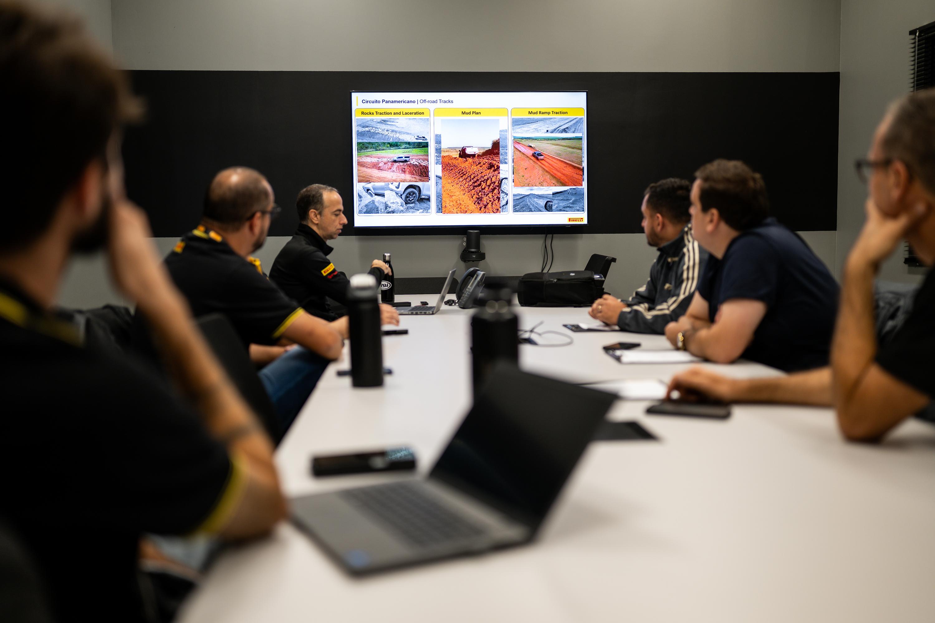 Homens em reunião, sentados em volta de uma mesa, observam uma apresentação em tela grande com imagens de máquinas em canteiros de obras