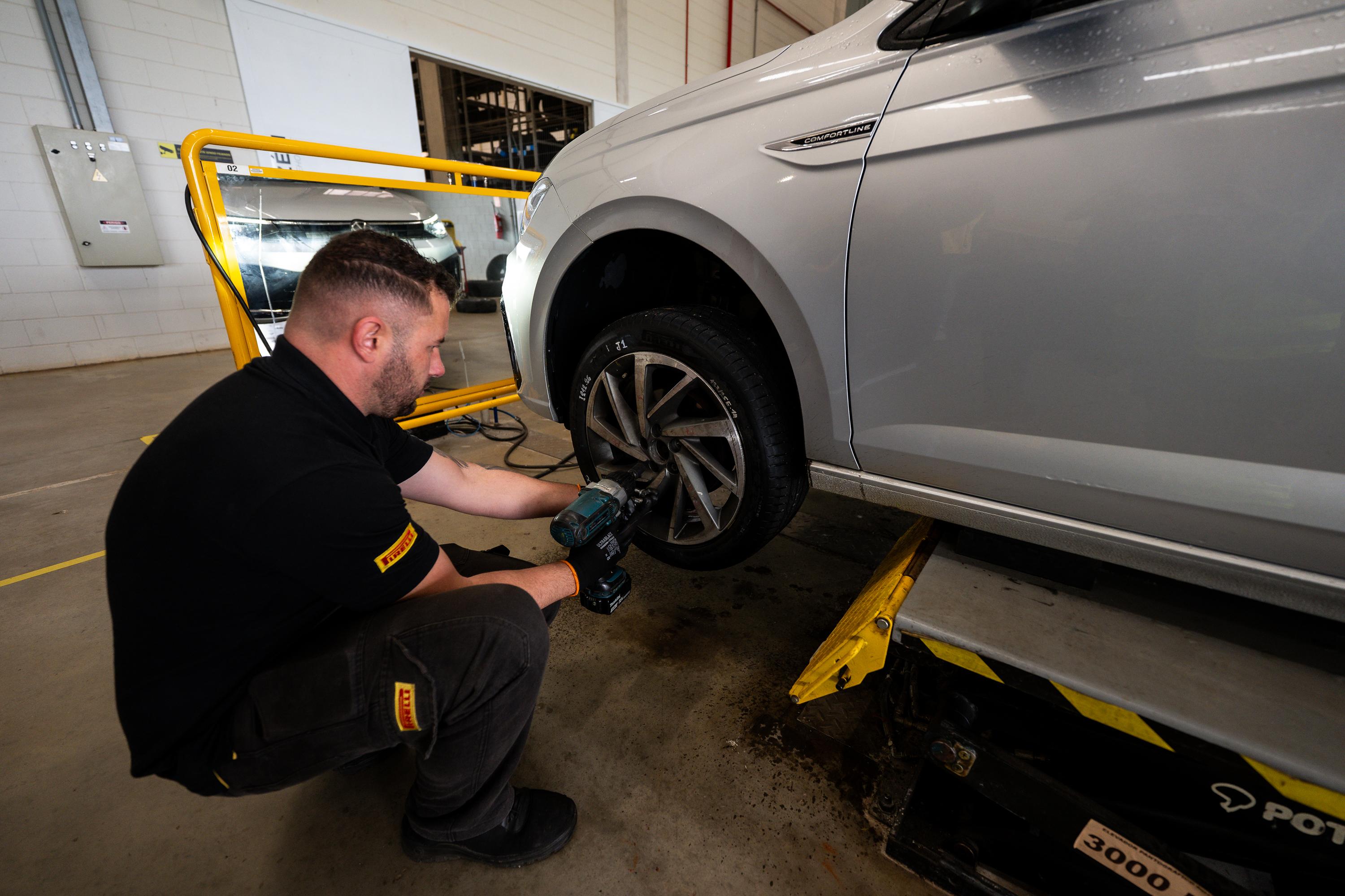 Mecânico barbudo, de camisa polo preta e calça escura com detalhes amarelos, agachado, usando uma parafusadeira elétrica para remover a roda dianteira de um carro prata, que está suspenso por um elevador automotivo amarelo e preto em uma oficina