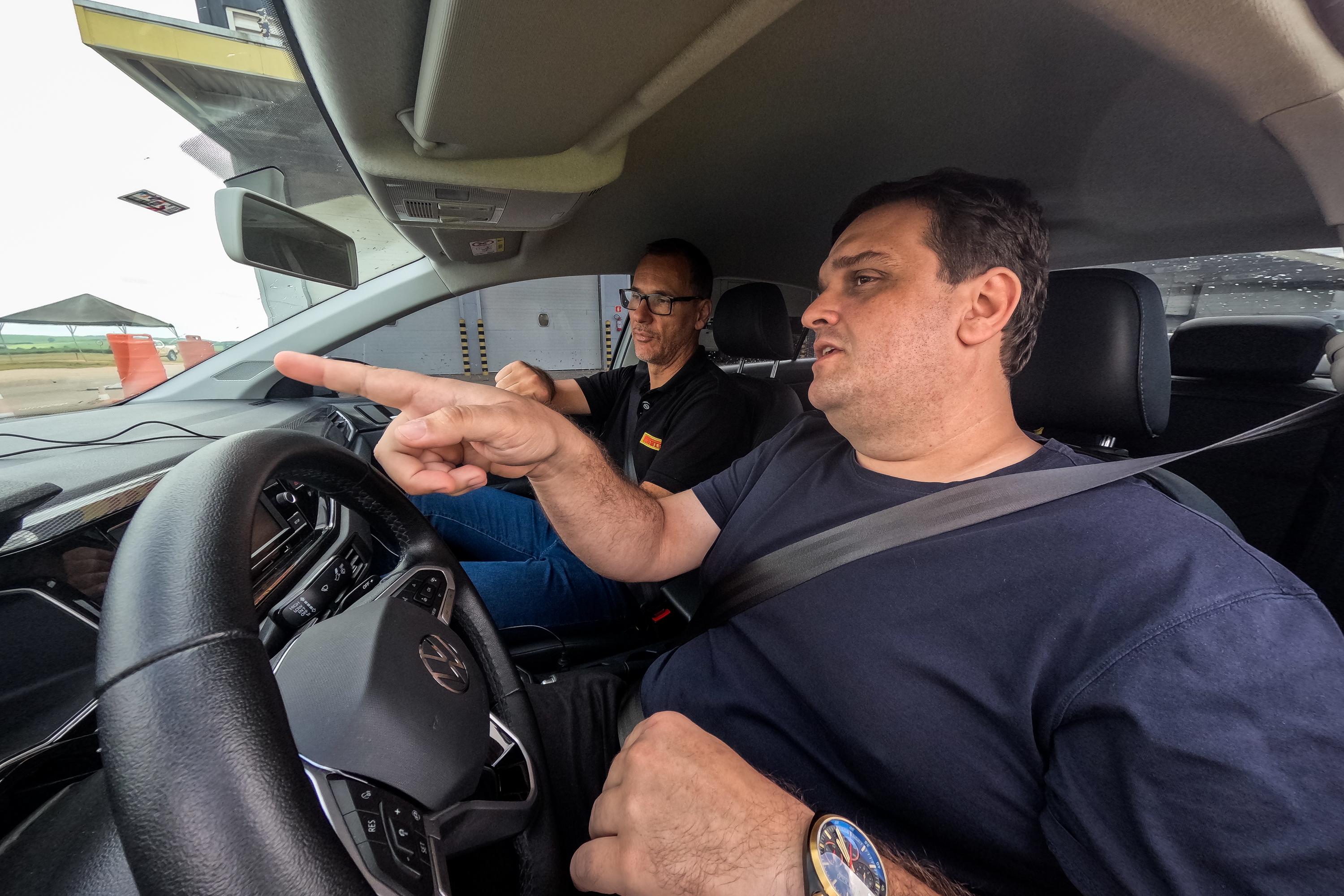 Homem de camiseta azul-marinho e cinto de segurança aponta para frente enquanto outro homem de óculos e camiseta preta dirige o carro