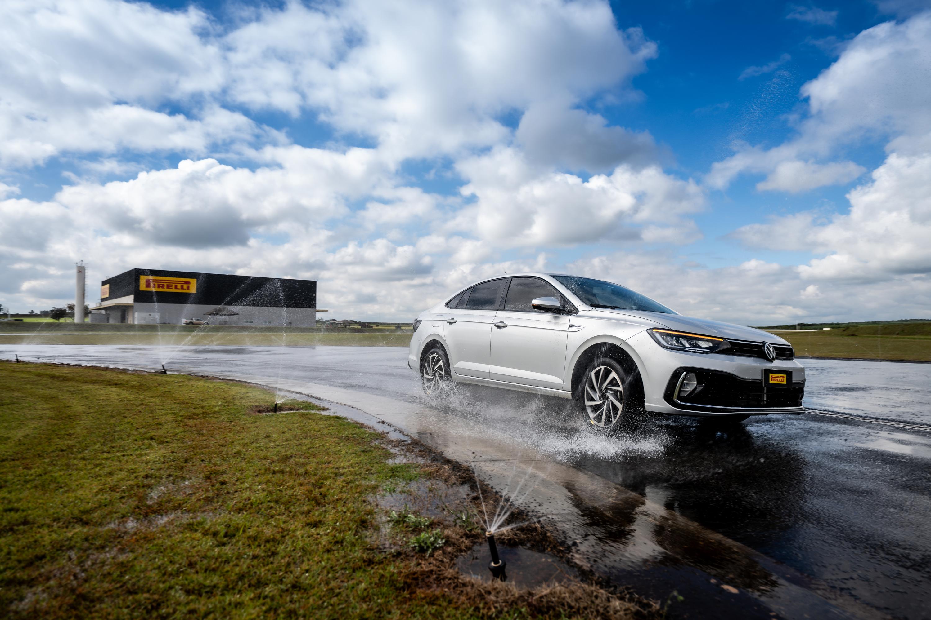 Carro sedan prata em pista molhada, com água espirrando dos pneus, sob céu azul com nuvens. Ao fundo, prédio escuro com logo Pirelli amarelo e branco.