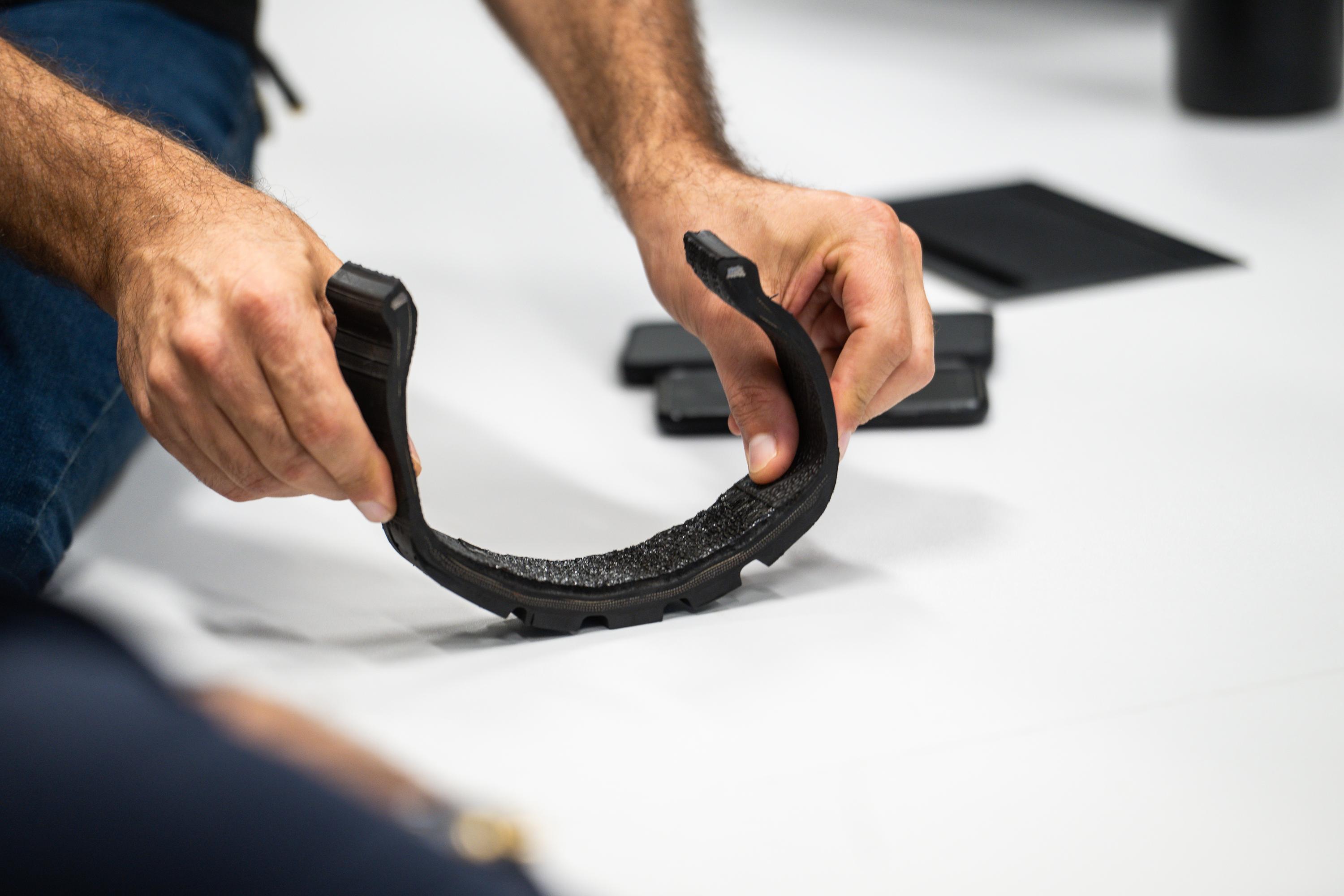 Mãos masculinas seguram e flexionam um objeto preto em forma de U, com textura rugosa na parte interna, sobre uma superfície branca