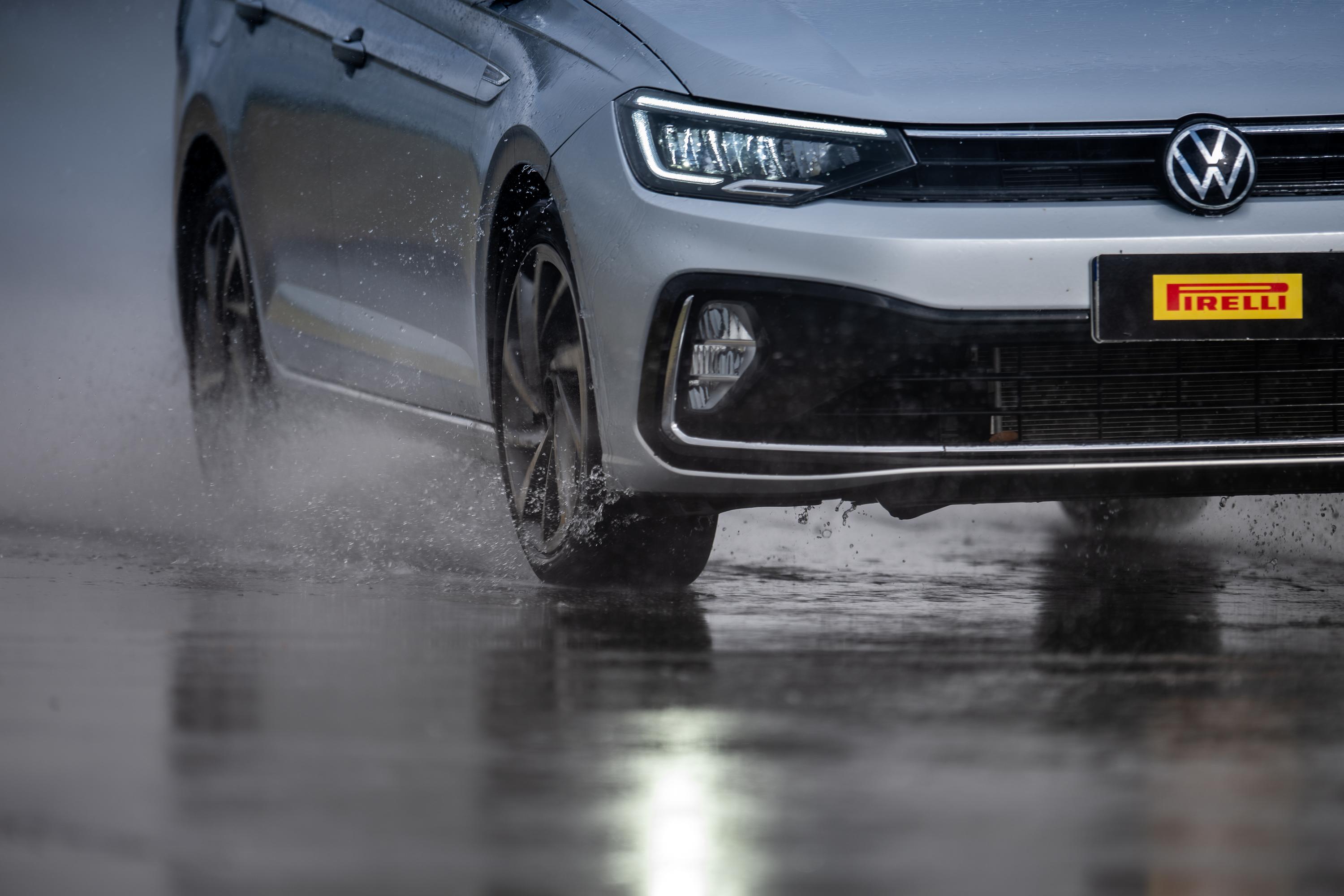 Carro cinza claro em movimento, com pneus levantando água em uma pista molhada. A frente do veículo exibe o logotipo da Volkswagen e uma placa preta com o nome PIRELLI em amarelo e vermelho