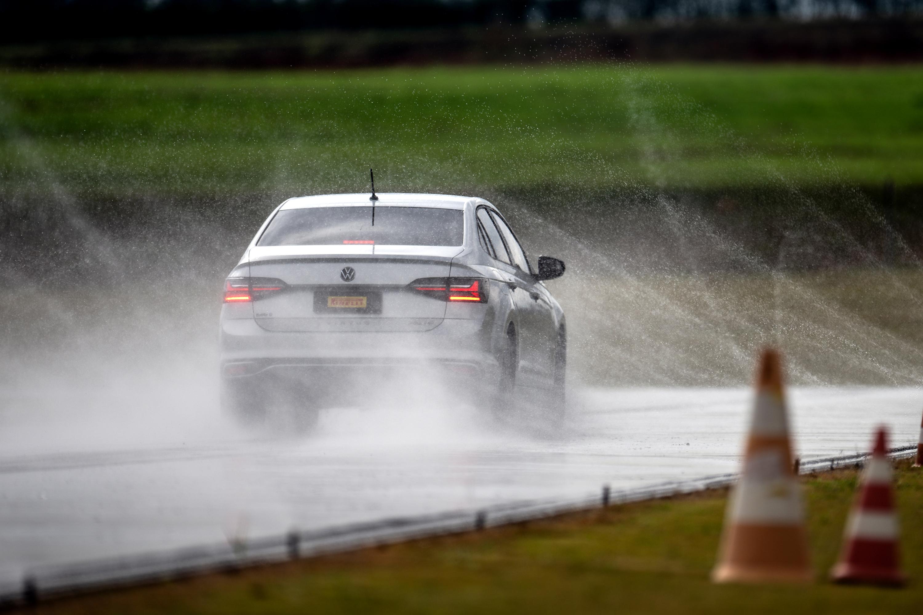 Carro sedan branco visto por trás, em pista molhada, levantando spray de água. Ao fundo, gramado verde e céu nublado. Cones de sinalização desfocados no canto inferior direito