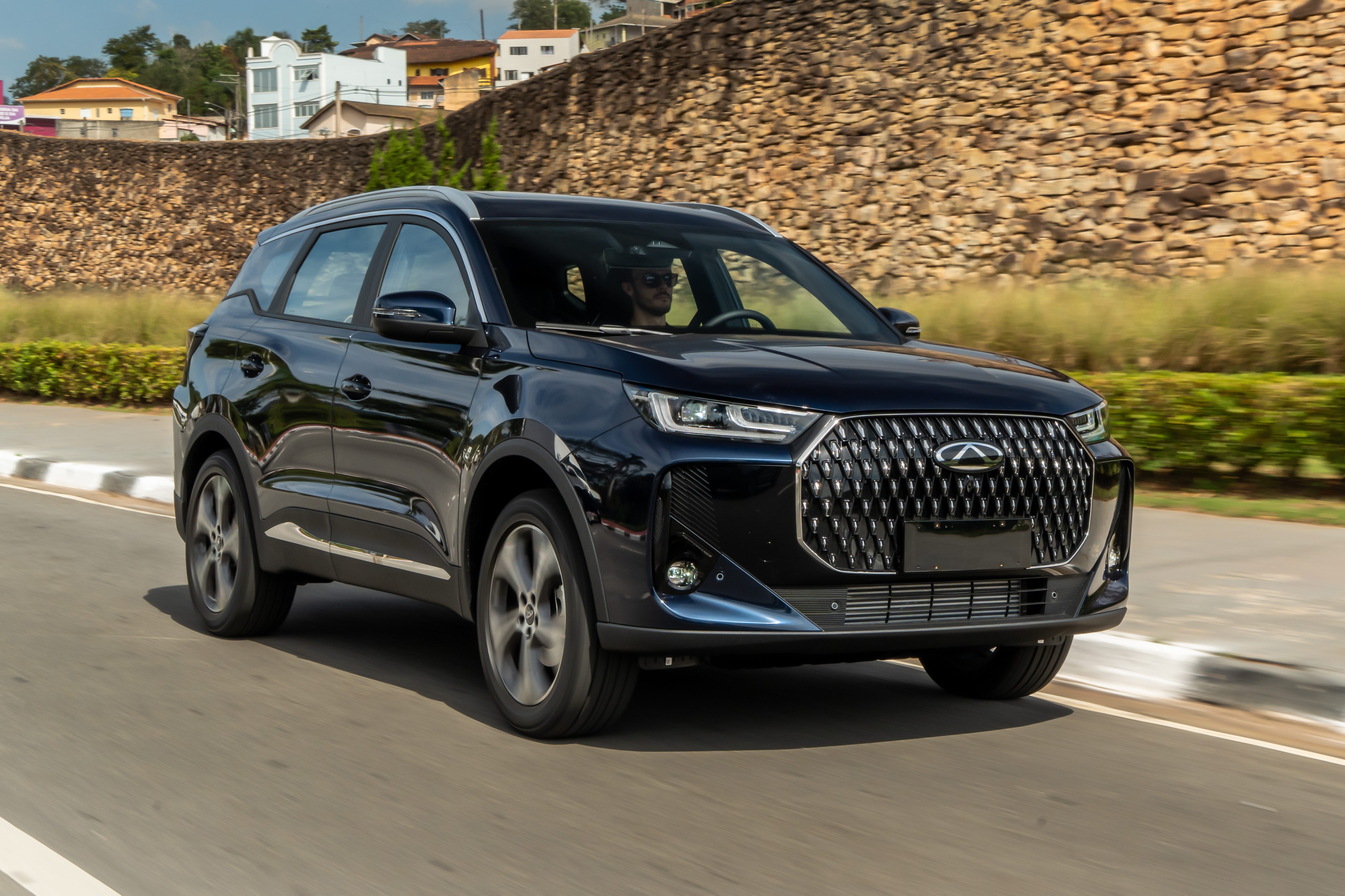 SUV preto brilhante em movimento na estrada, com um motorista usando óculos de sol. Ao fundo, um muro de pedras, casas e vegetação sob céu azul com nuvens