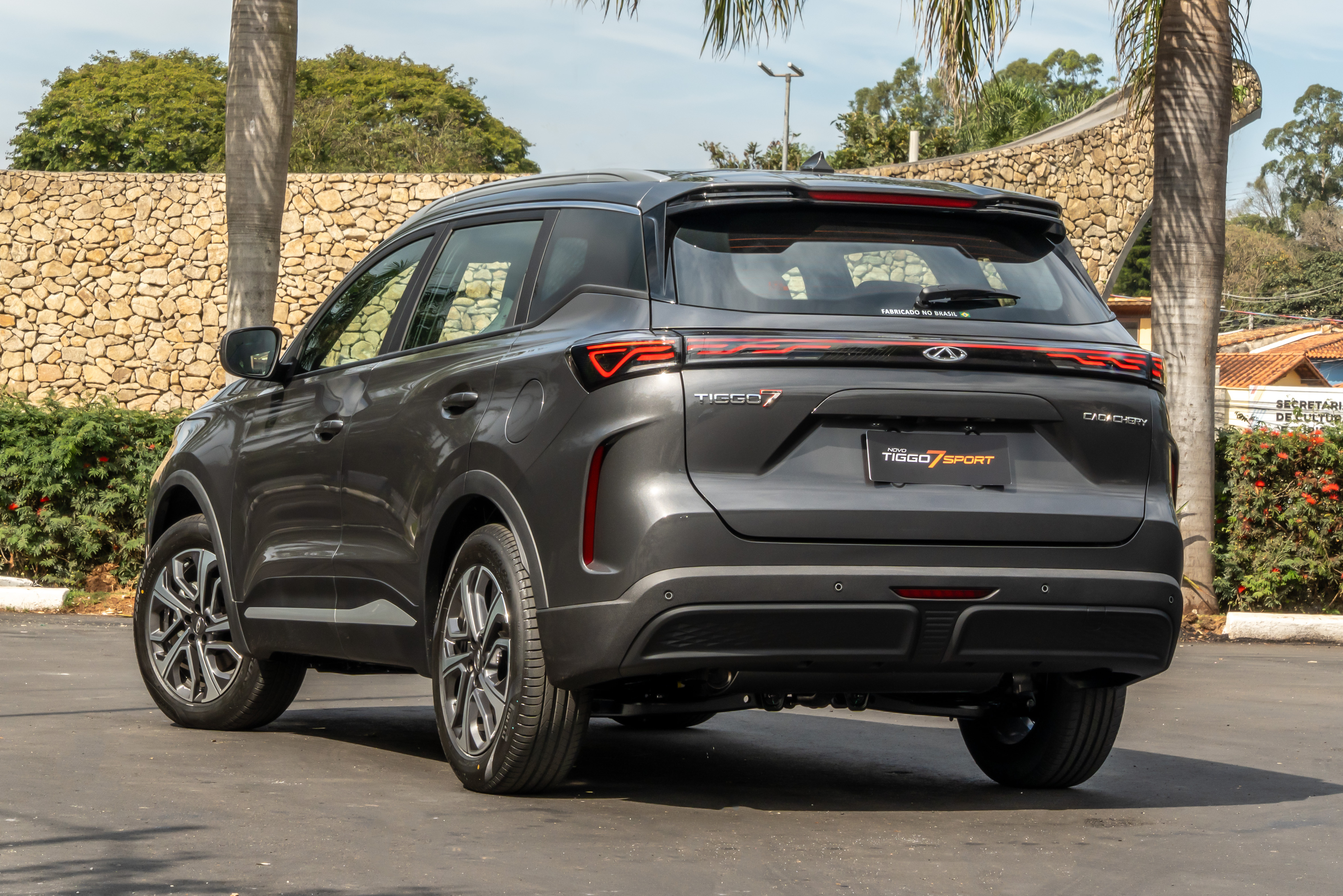 SUV Chery Tiggo 7 Sport cinza escuro, visto de traseira e lateral, estacionado em pátio asfaltado com muro de pedras e palmeiras ao fundo