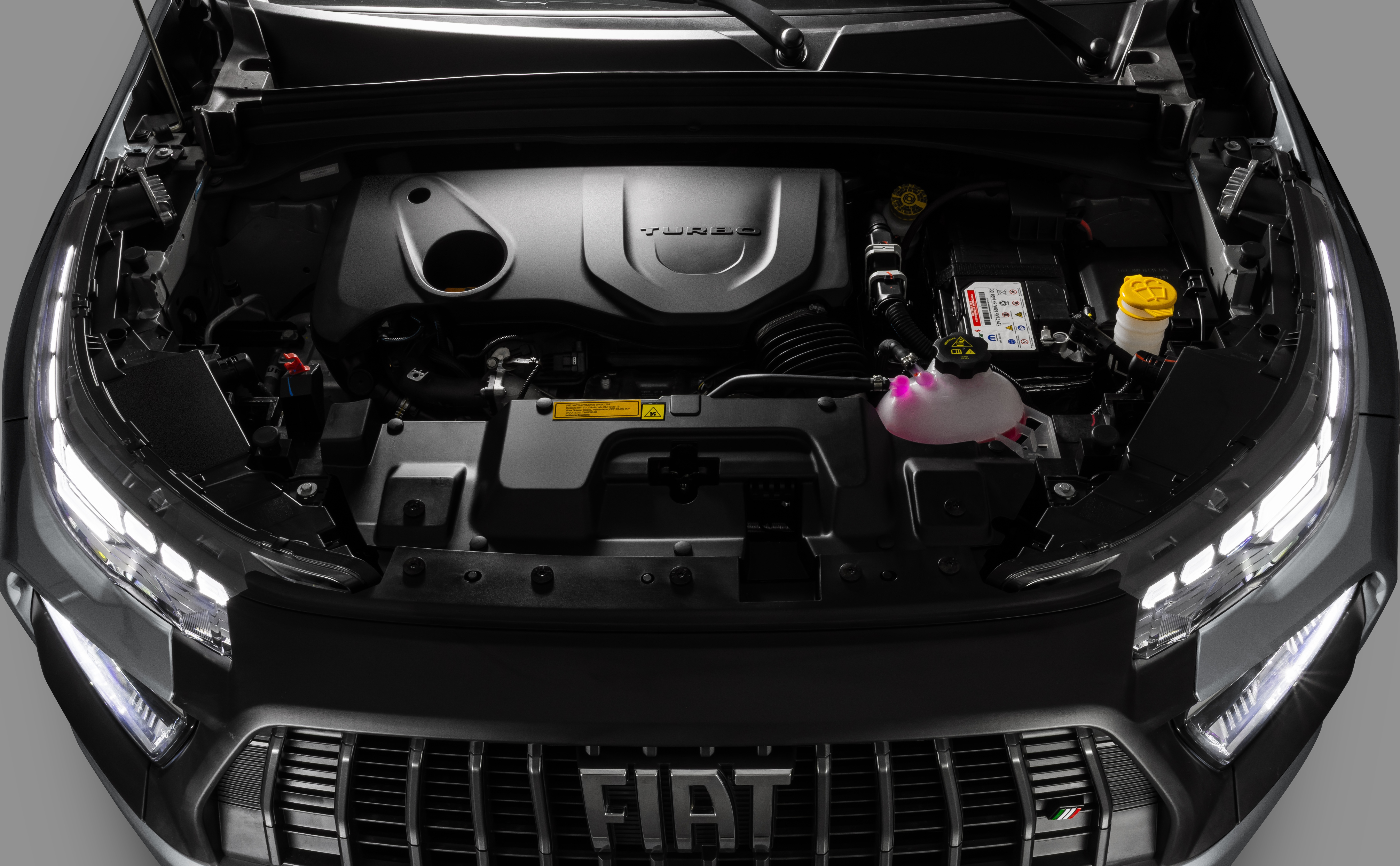 Motor de carro preto com capô aberto, revelando o motor turbo, reservatório de fluido rosa e bateria. A grade frontal com o logo Fiat é visível abaixo, com faróis de LED acesos
