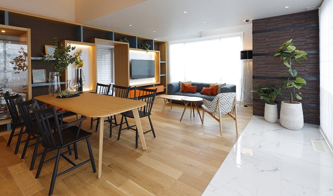 Sala de estar e jantar com mesa de madeira clara e cadeiras pretas, sofá cinza com almofadas laranja, estante com TV e plantas, e parede de tijolos escuros com vasos brancos