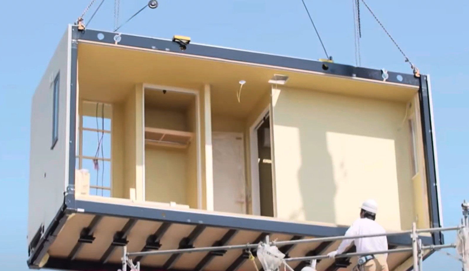 Um módulo de casa pré-fabricada, com paredes internas claras e janelas, sendo içado por guindaste. Um trabalhador de capacete branco e camisa clara observa a operação de uma estrutura de andaimes abaixo