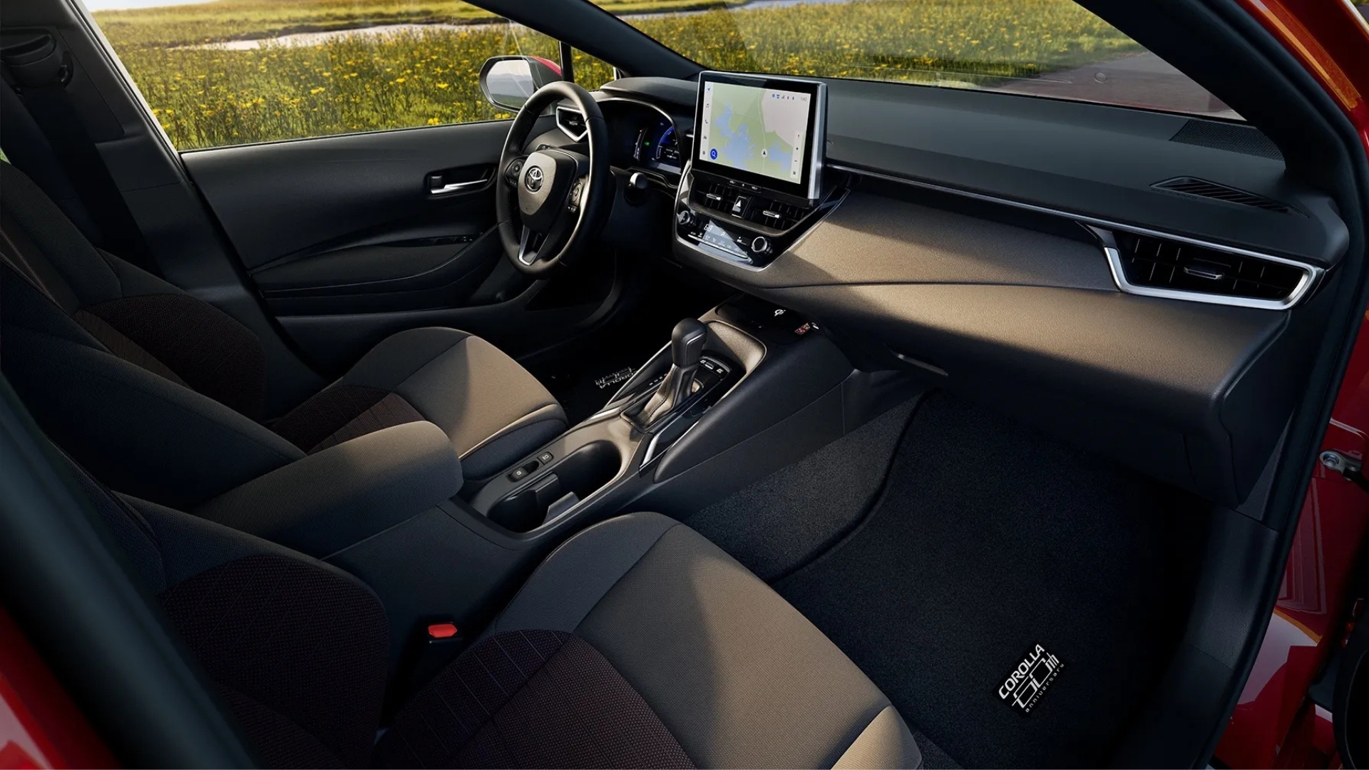Interior de um carro moderno, com bancos de couro escuro e detalhes em cinza. O painel possui uma tela multimídia com mapa, volante multifuncional e câmbio automático. A luz do sol entra pela janela, iluminando o campo verde ao fundo