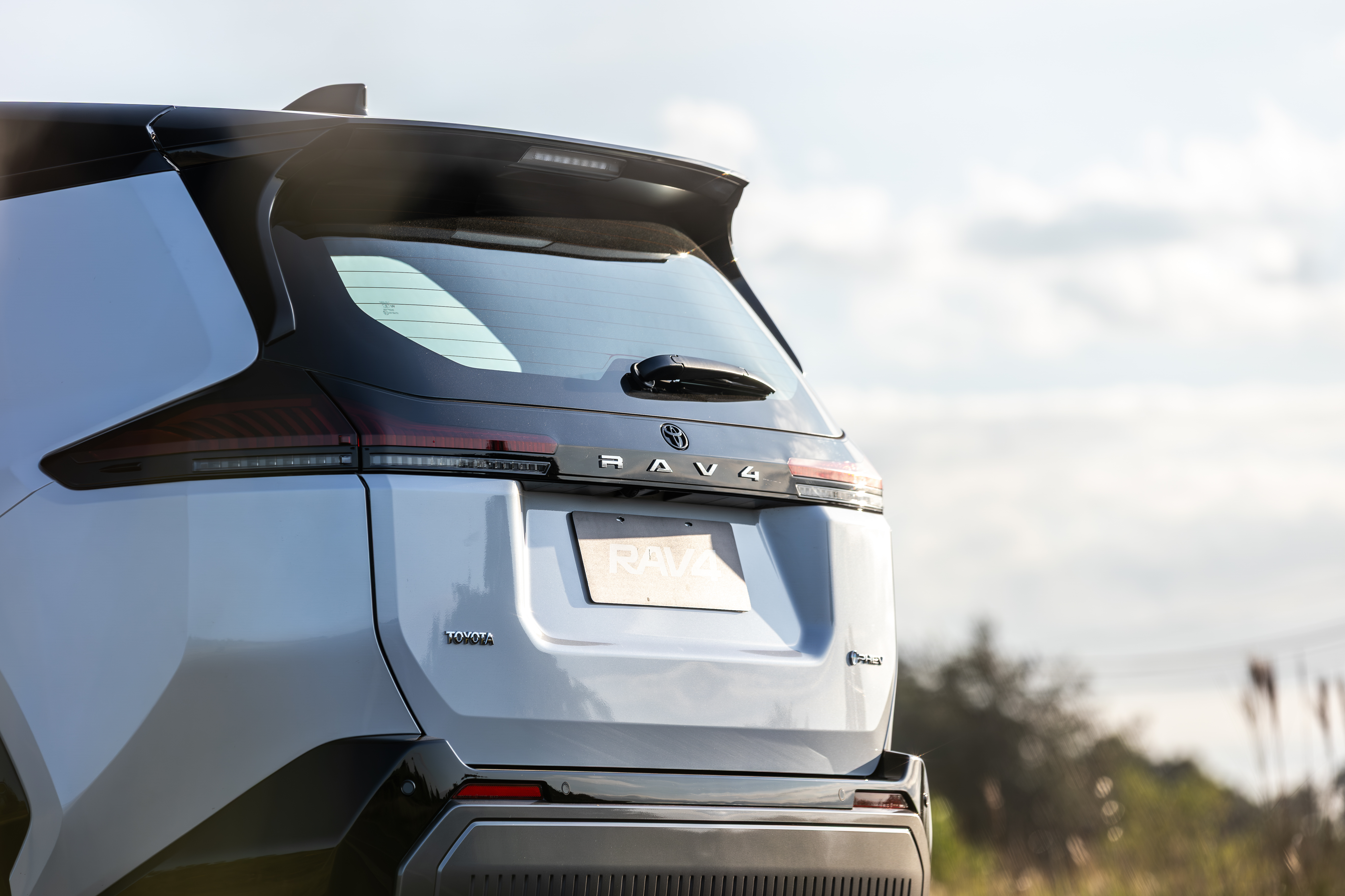 Traseira de um SUV Toyota RAV4 cinza claro, com lanternas horizontais escuras e aerofólio preto, sob céu azul com nuvens esparsas