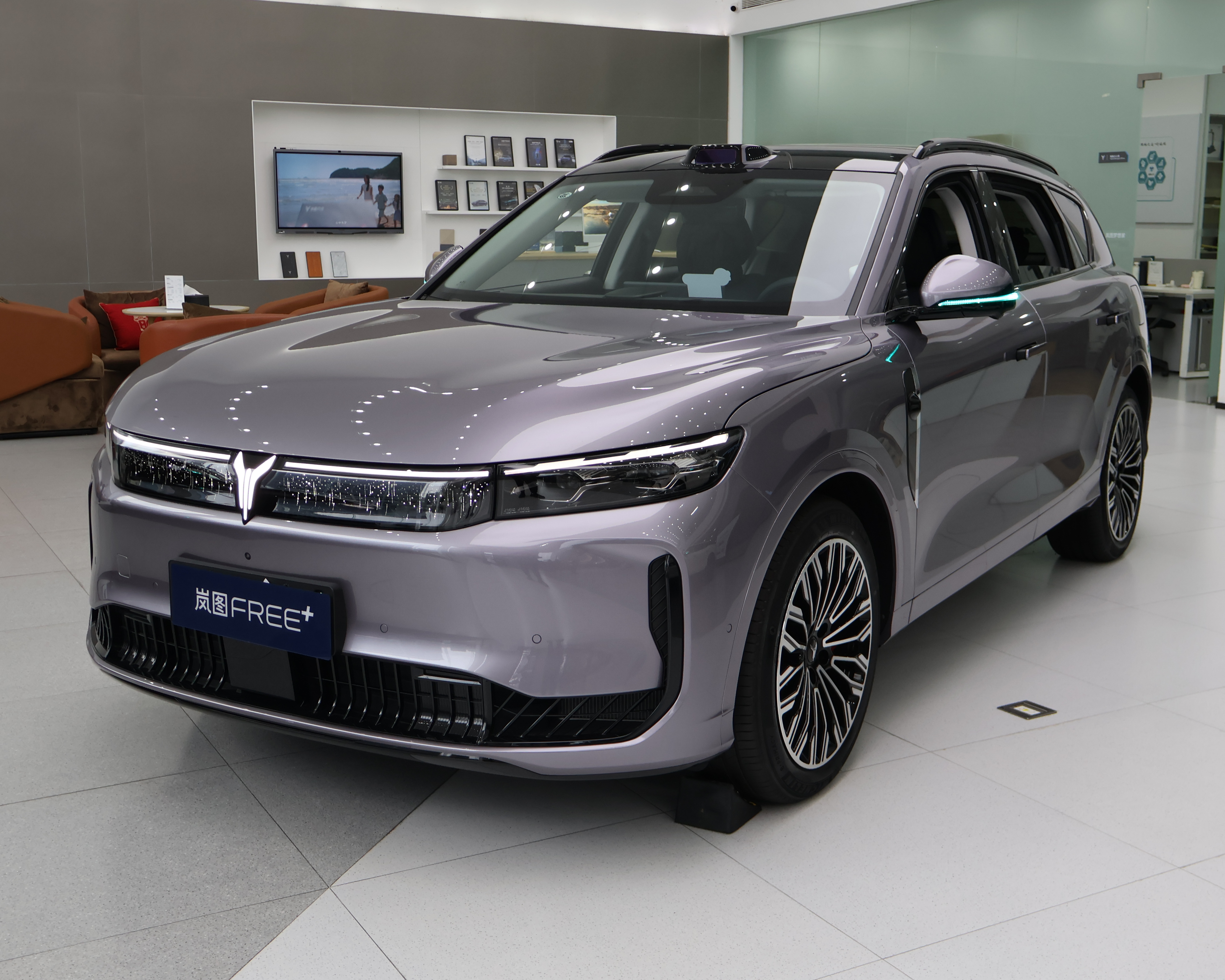 SUV cinza-claro com design moderno, faróis finos e grade frontal preta, exibido em showroom com piso claro e área de estar ao fundo