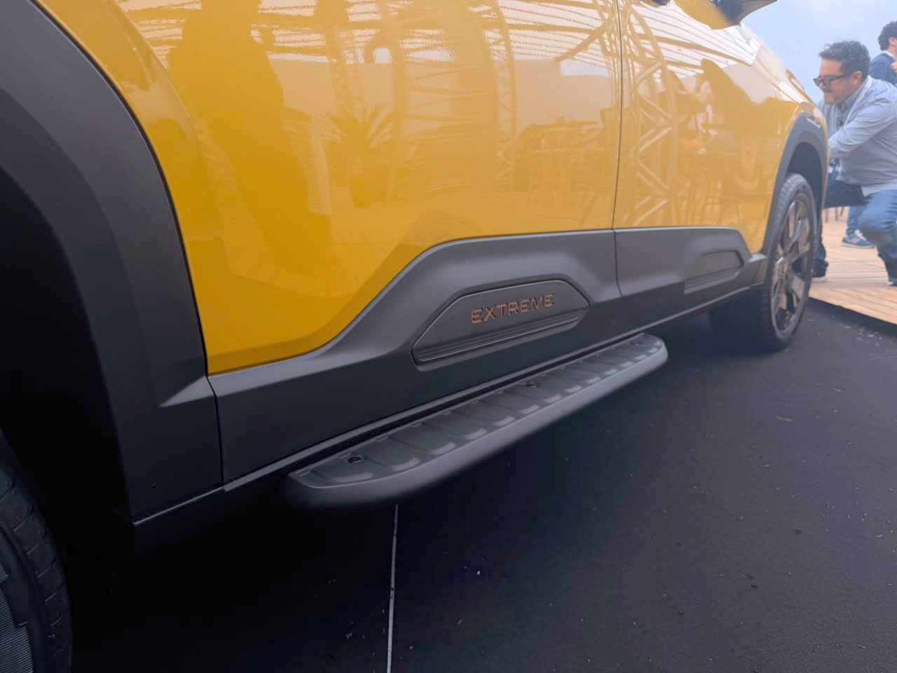 Lateral de um carro amarelo com detalhes em plástico preto, incluindo um estribo e a inscrição EXTREME em laranja. Um homem de óculos escuros e camisa clara observa o veículo ao fundo