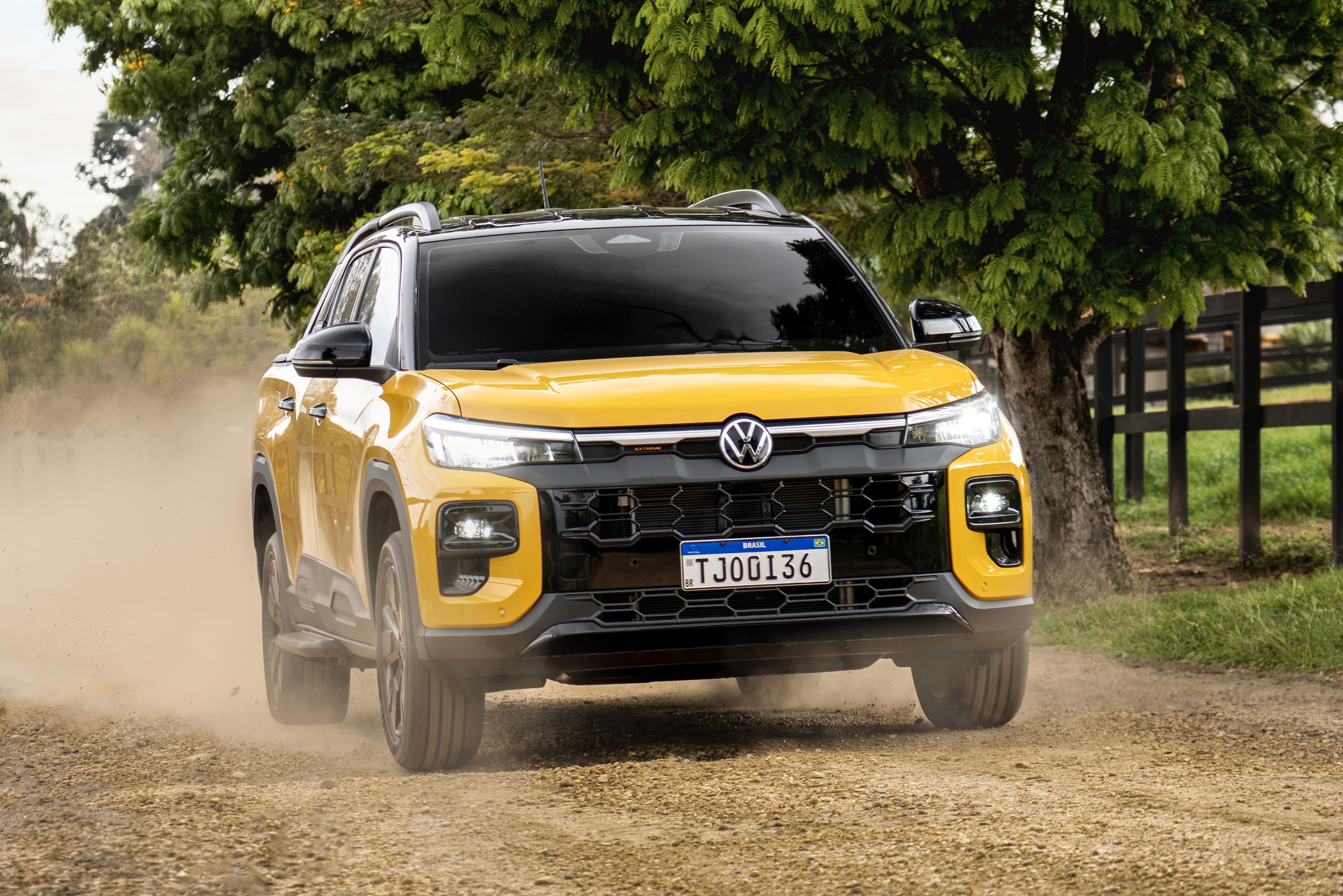 Volkswagen Tukan amarelo brilhante, com teto preto e rack de teto prateado, em estrada de terra levantando poeira. Ao fundo, árvores verdes e uma cerca de madeira escura. A placa do carro é TJQ0136