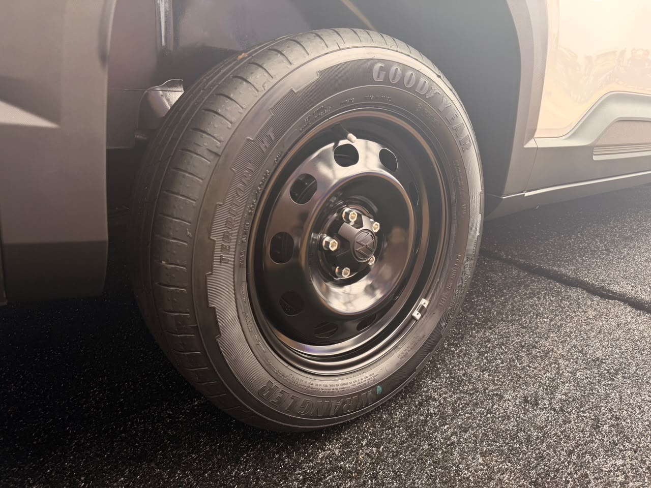 Pneu Goodyear Territory HT preto com aro de ferro e cinco parafusos dourados, montado em um carro escuro, estacionado em asfalto cinza com reflexo de luz solar