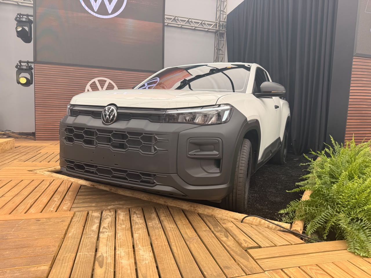 Picape branca com para-choque frontal cinza escuro em destaque, estacionada sobre uma rampa de madeira. Ao fundo, um painel com o logo da Volkswagen e cortinas pretas. À direita, folhagens verdes.