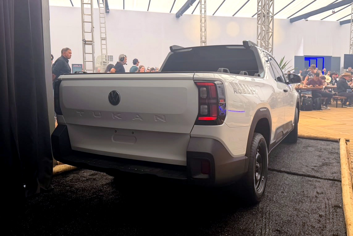 Traseira de uma picape branca Volkswagen Tukán, com lanternas verticais acesas e o logo da marca no centro. Pessoas conversam ao fundo, em um ambiente interno com iluminação clara e estrutura metálica no teto