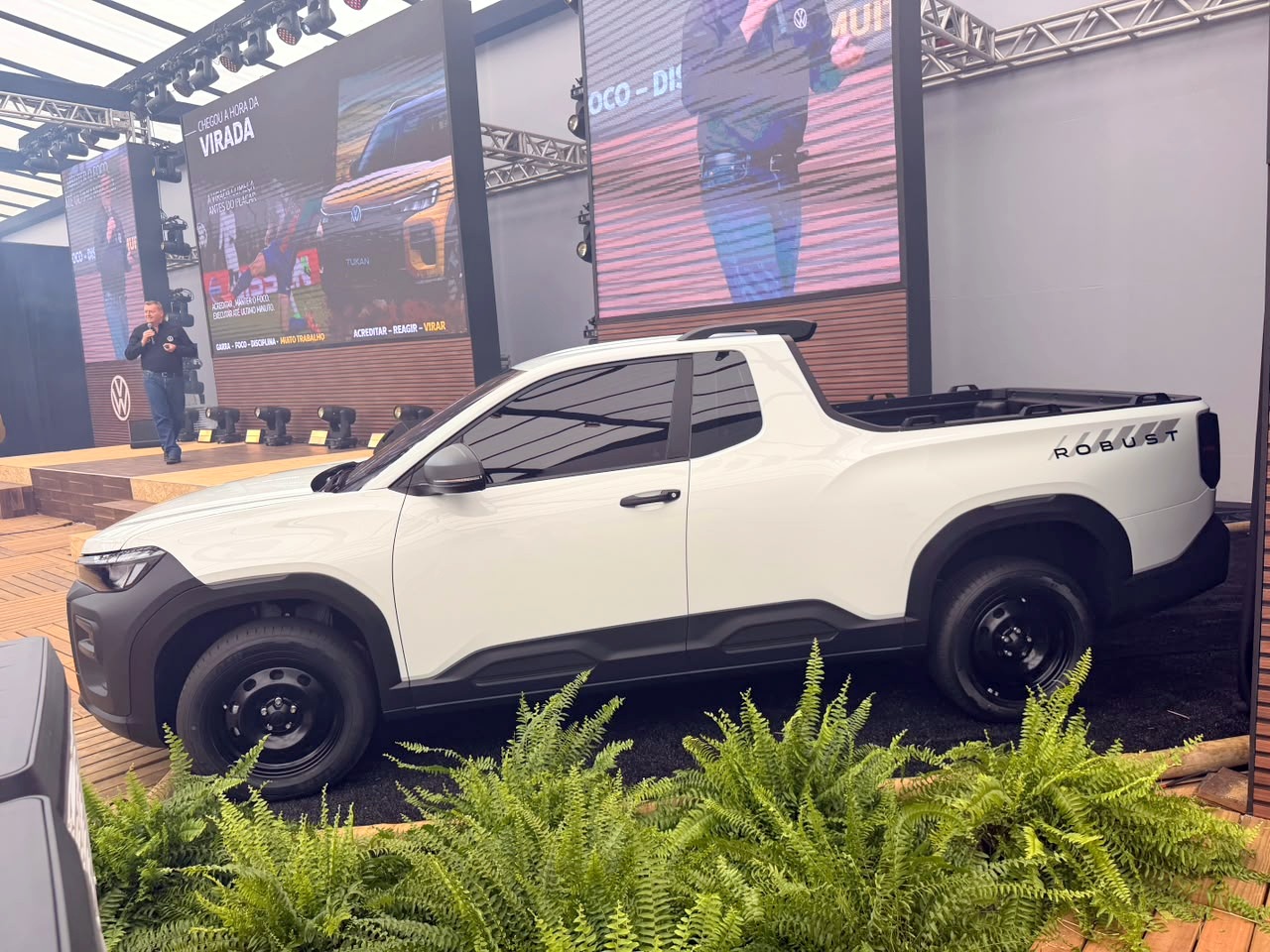 Picape branca de cabine simples, modelo Robust, estacionada em um palco com folhagens verdes na frente e telões exibindo carros e pessoas ao fundo