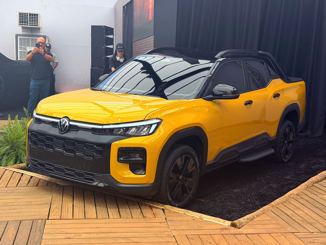 Picape Volkswagen amarela com teto preto, exibida em um palco de madeira. Ao fundo, um homem fotografa e uma mulher usa fones de ouvido
