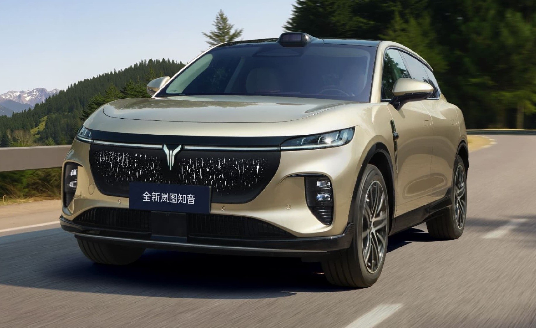 SUV dourado em movimento numa estrada sinuosa, com montanhas e árvores verdes ao fundo. O carro tem uma grade frontal preta com um emblema prateado e um sensor preto no teto. Na placa frontal, lê-se 全新岚图知音